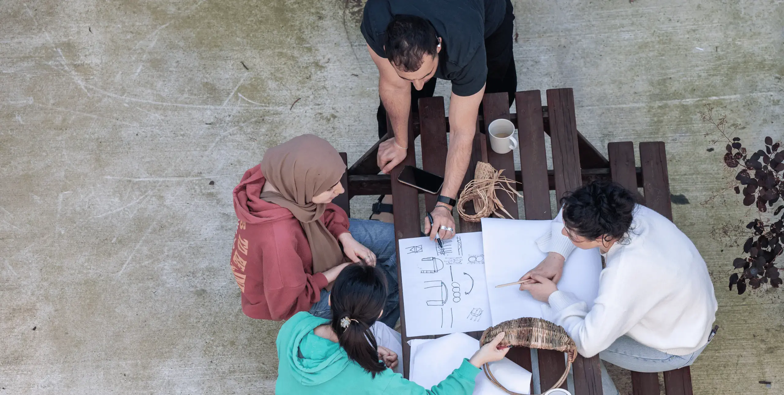 An overhead shot of a group of four people sitting and standing around a wooden picnic bench, looking at papers and crafting materials spread on its surface, seemingly in conversation and working on something together.