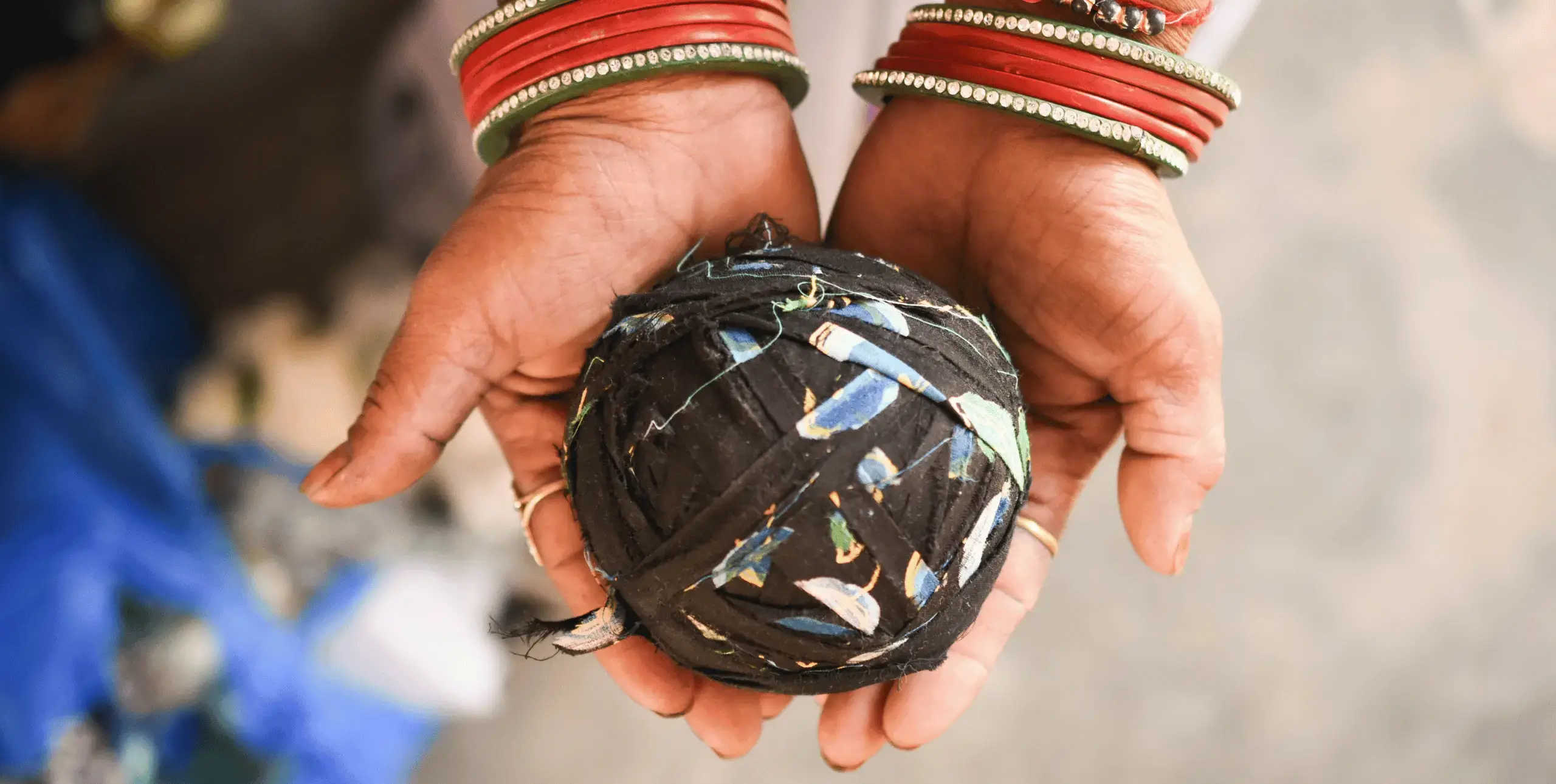 A close-up of a woman's hands holding a tightly wound ball of fabric. The woman wears brightly coloured bangles on her wrists.