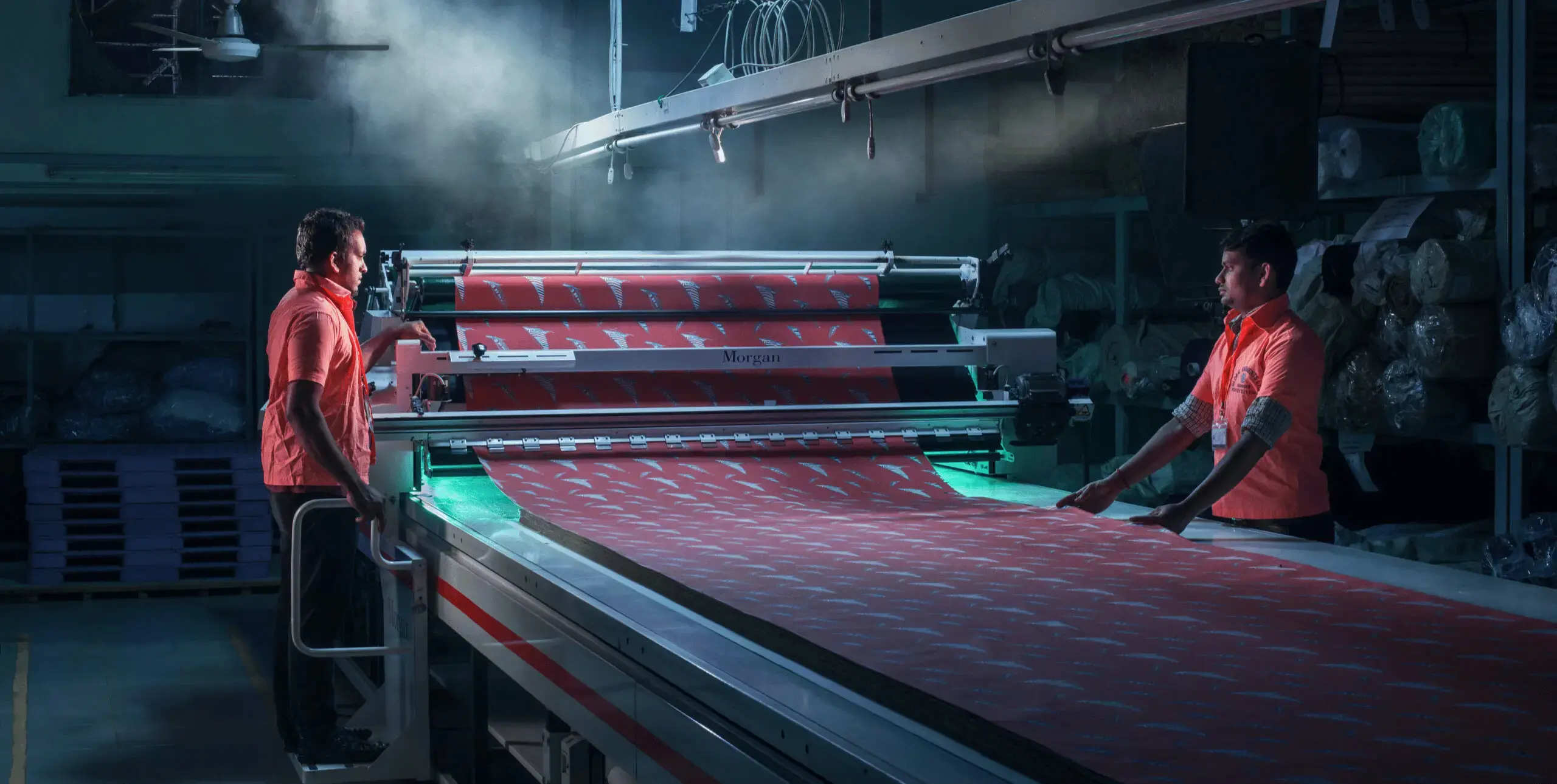 Two men stand either side of a fabric production line in a factory.