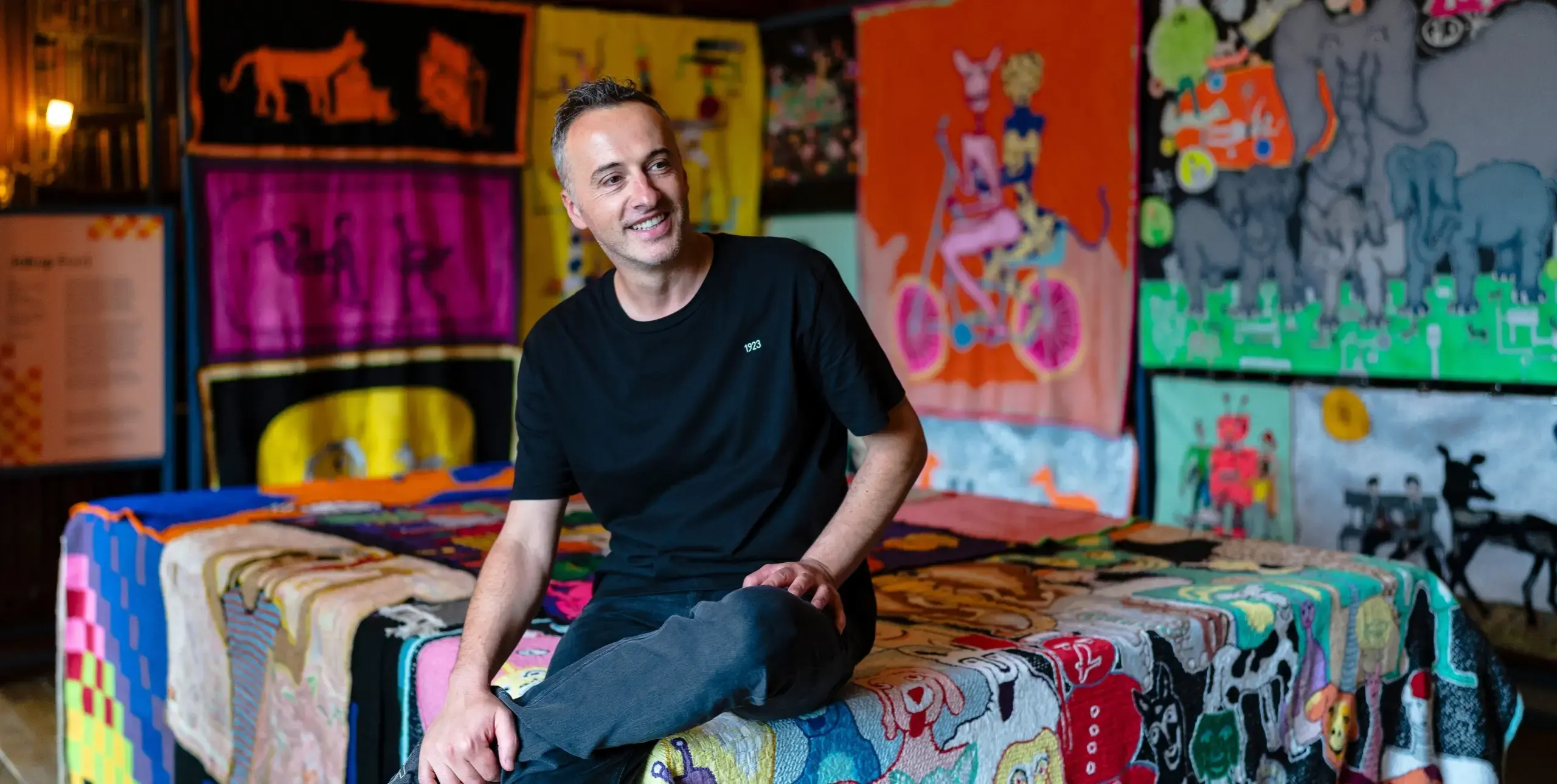 Man smiling, sat on fabric artwork with artwork around him.