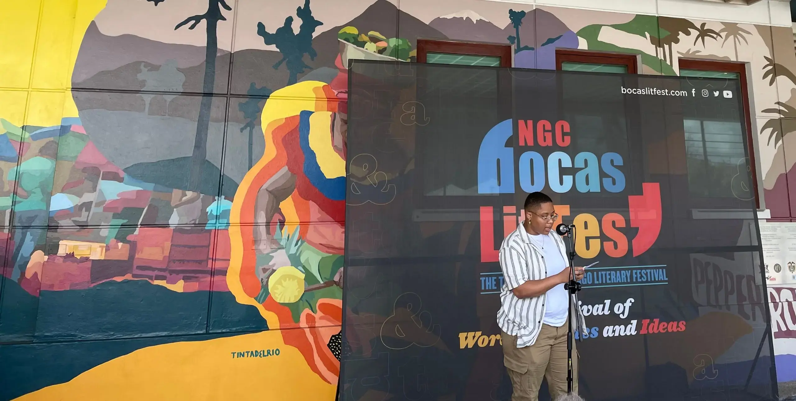 A writer speaks in front of a colourful mural and Bocas Lit Fest banner