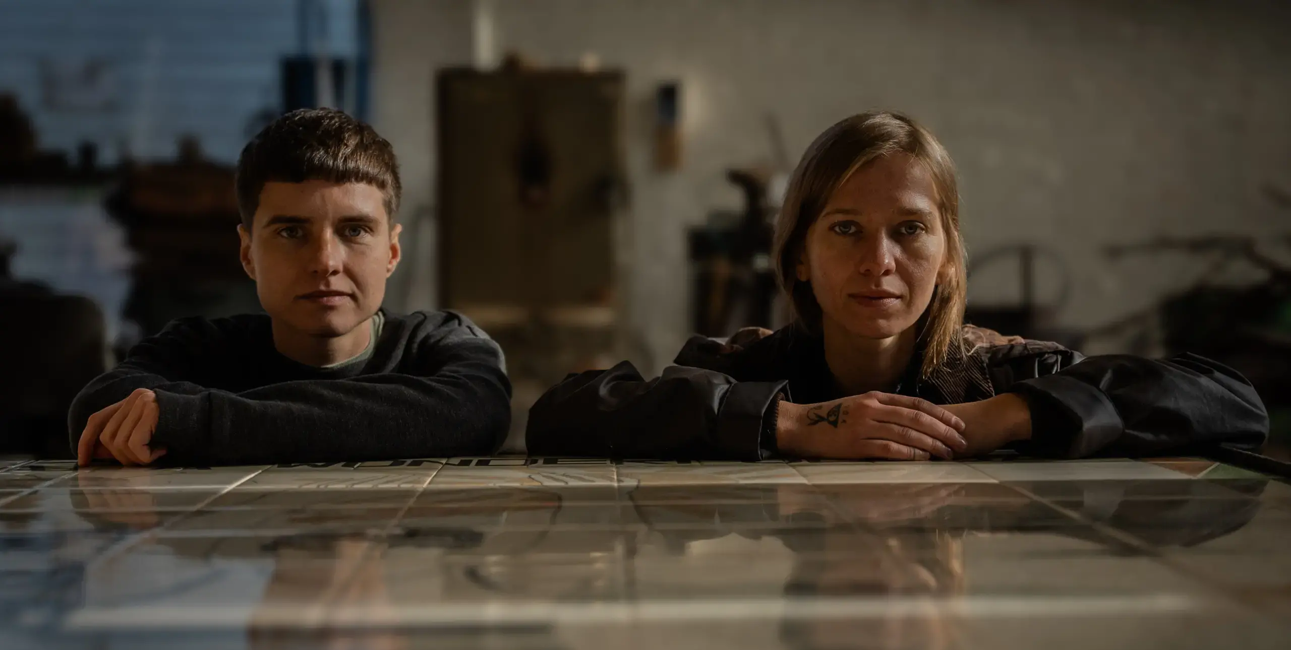 Two people sit alongside each other with their arms resting on a shiny tabletop. They are looking straight ahead, into the camera.
