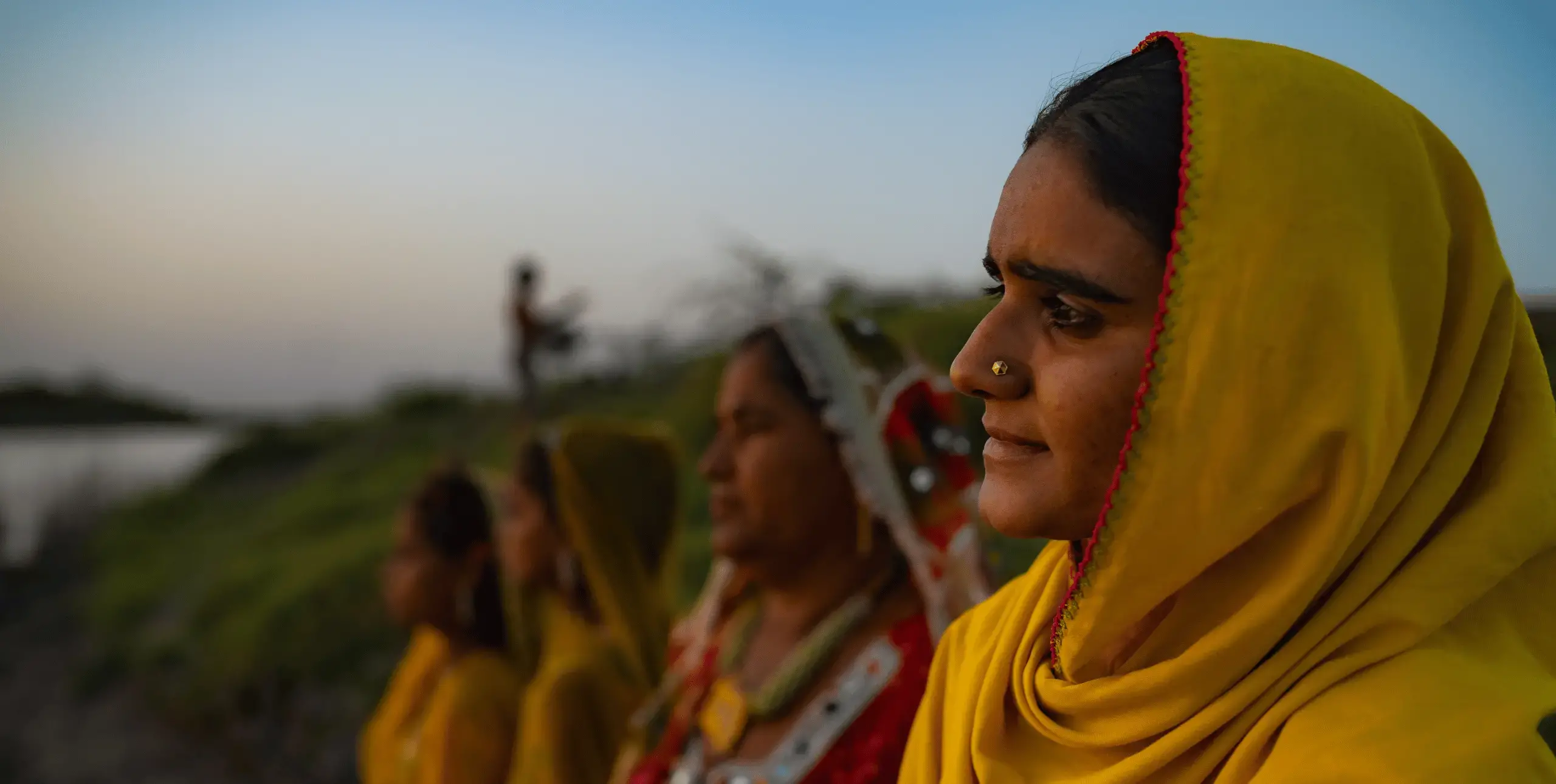 A line of four women stand in a line reaching diagonally left from the right hand side of the image. The woman closest to the camera is in focus and the rest are out of focus, as is the background, which appears to be a rural setting next to water. The women are wearing yellow head coverings.