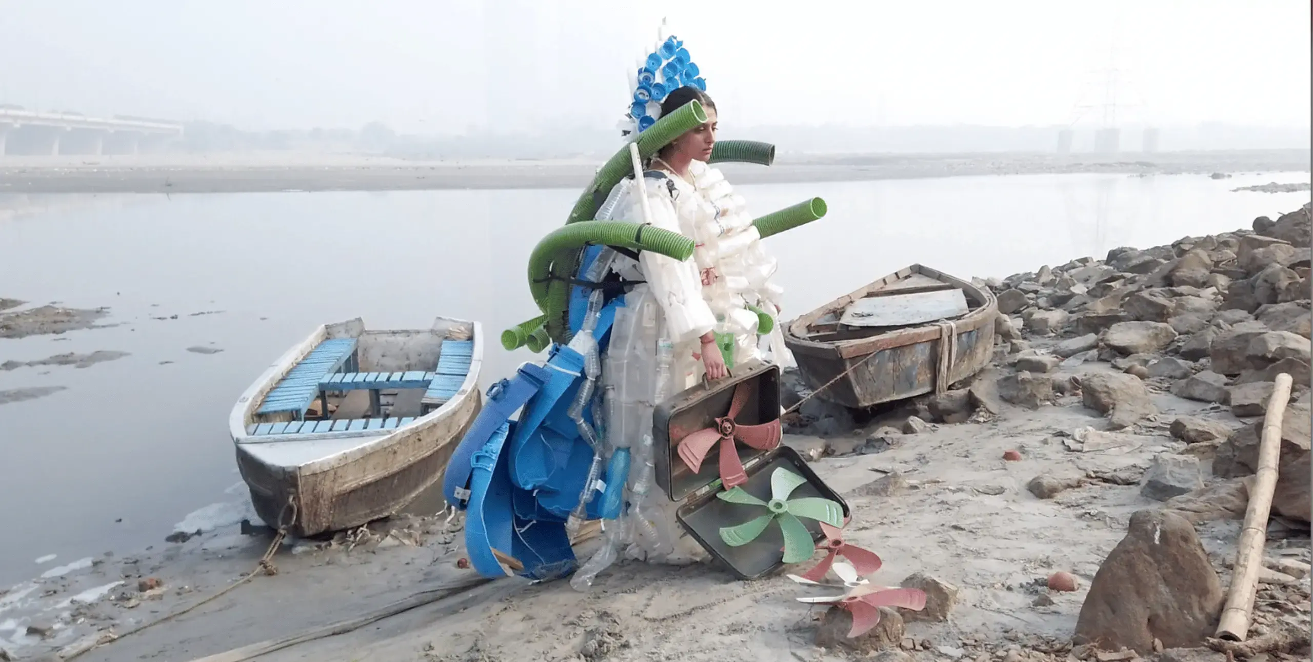 A woman sits on the banks of a wide river surrounded by abandoned boats and rocks. She wears a costume made from plastic bottles, tubing and machinery parts.