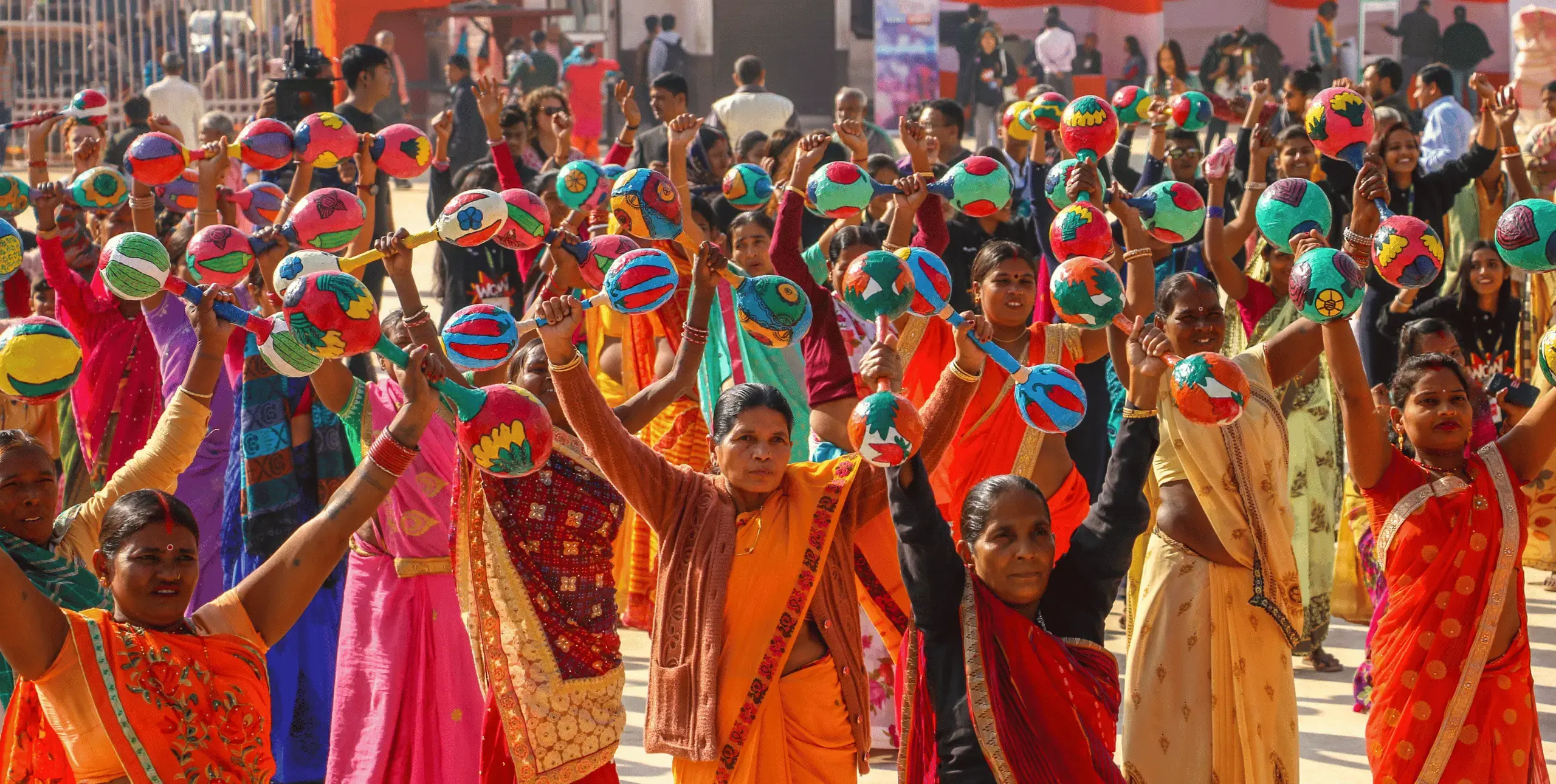 WOW Nepal, 2019. A crowd of women wearing brightly coloured saris stand with their arms raised holding what looks like large papier mache maracas.