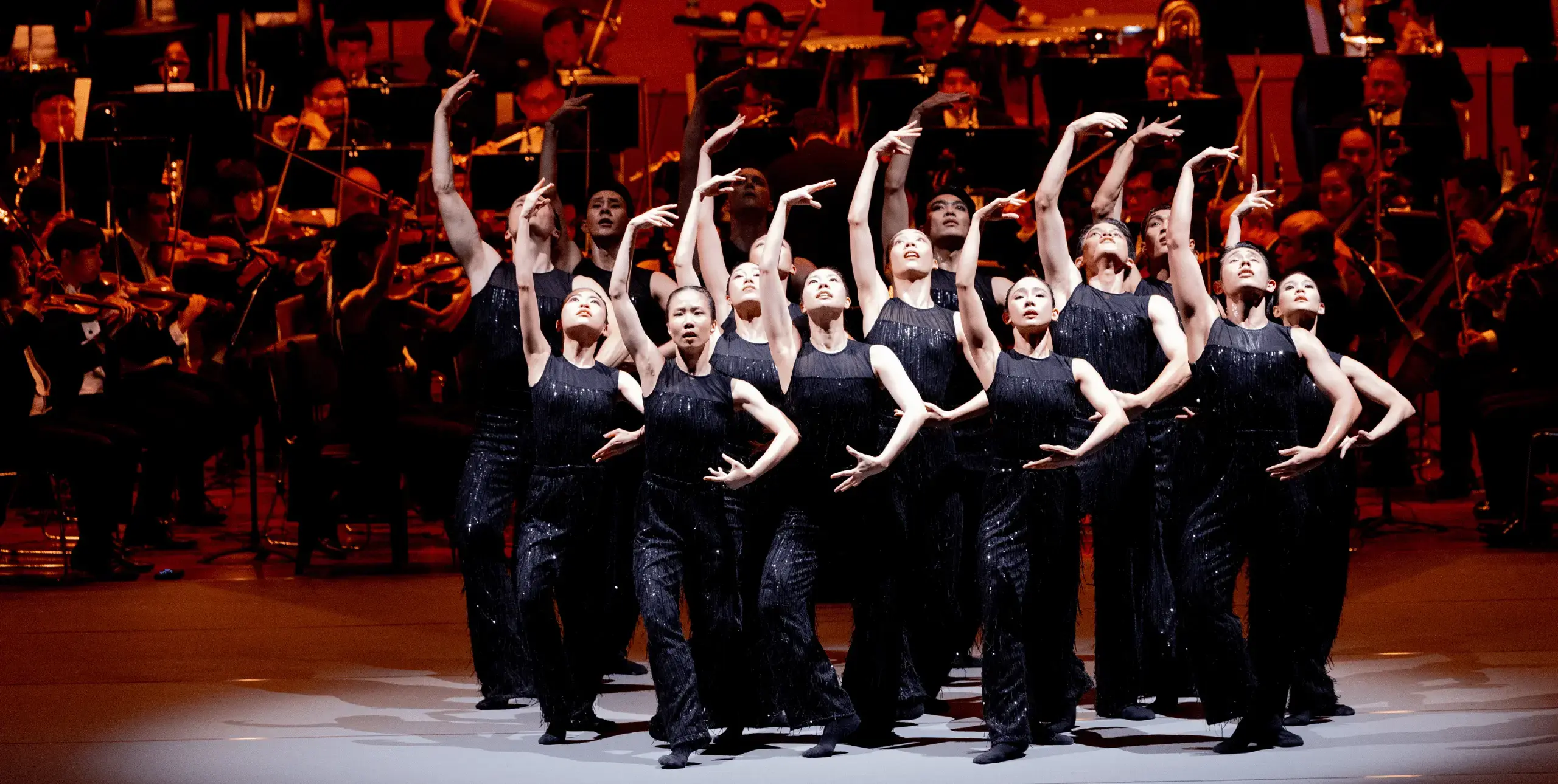 A group of dancers dressed in black stand in the same balletic pose on stage, with their right arms held above their heads, and their left arms below, looking upwards 