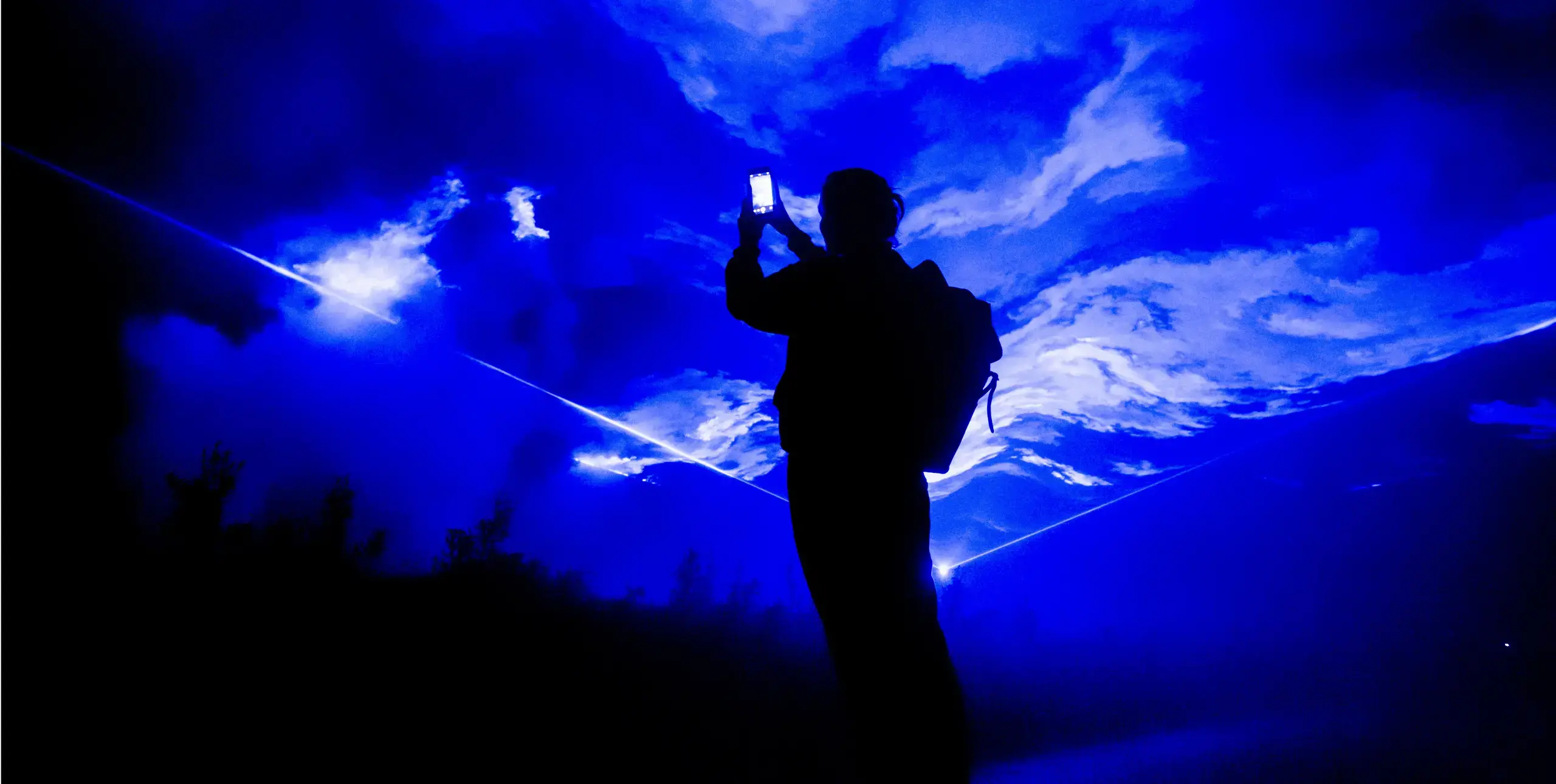 A figure stands silhouetted against a projection of bright blue and white swirling clouds. The figure holds up a mobile phone with a lit-up screen, as if taking a photo.