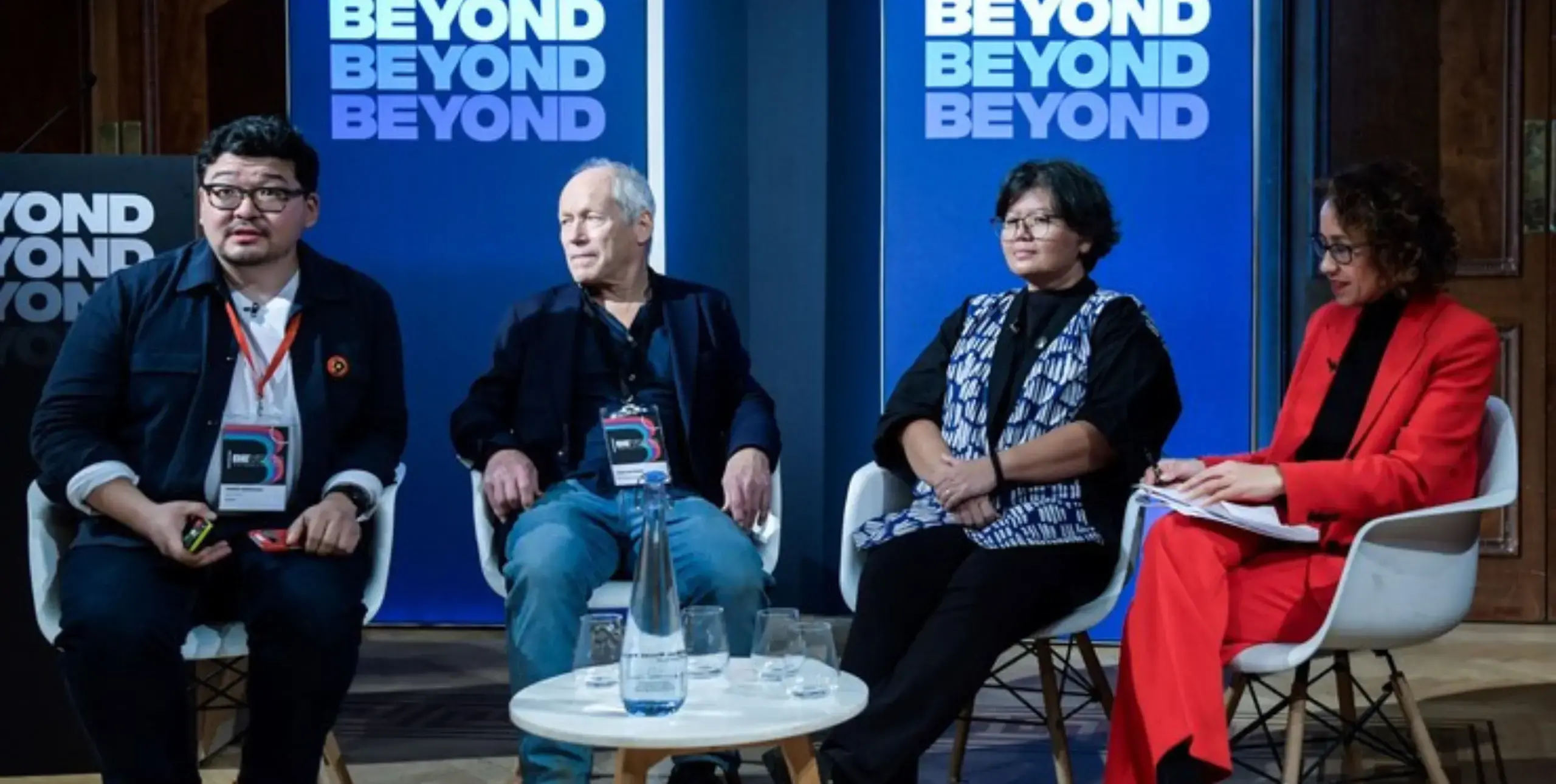 Four people seated on a stage in front of banners reading 'BEYOND' at the BEYOND conference 2023. The people are Daniar Amanaliev, John Newbigin, Tita Larasati and Samira Ahmed.