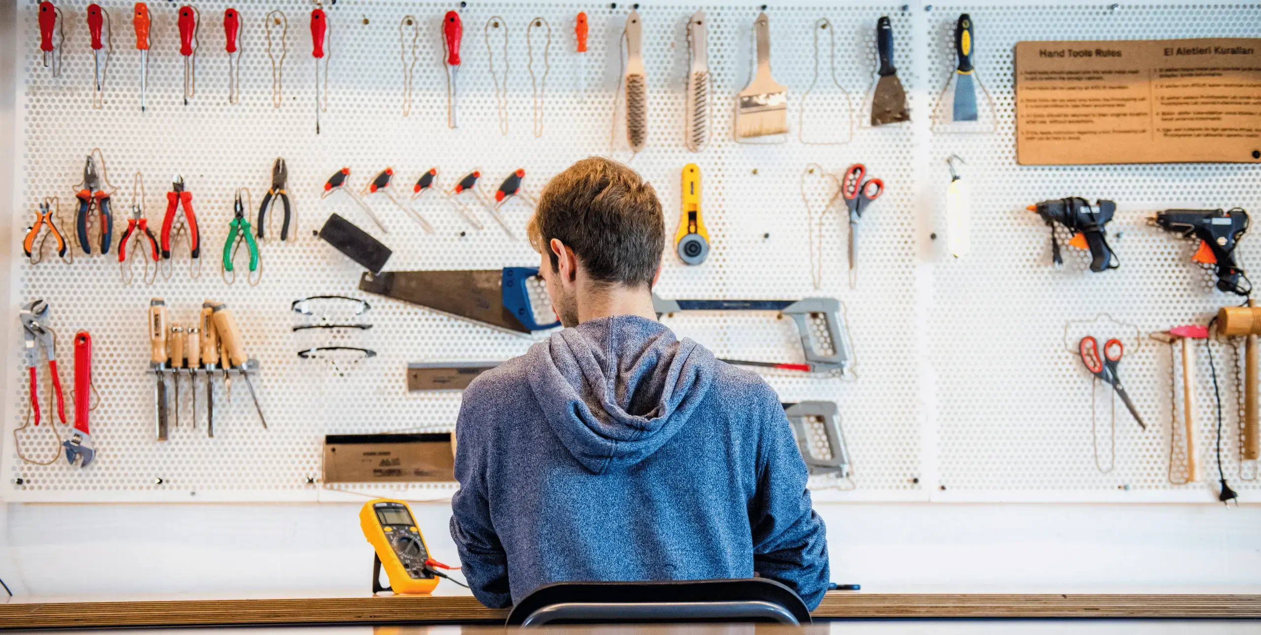 A man sits at a table or workbench with his back to the camera. In front of him is a wall with various tools hanging on it.