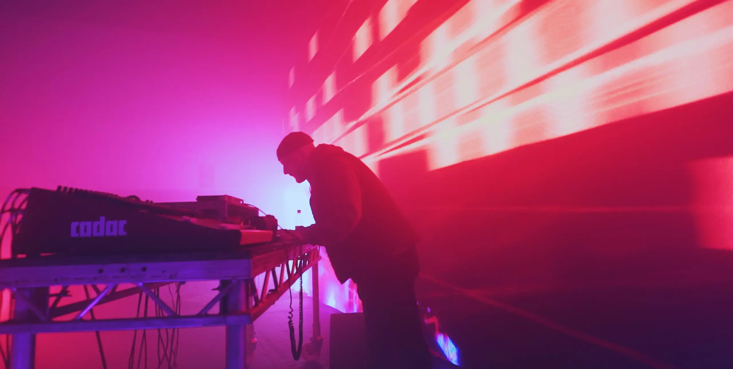 A DJ stands at a set of decks, lit from behind against a purple/pink and orange brightly lit backdrop. He leans forwards.