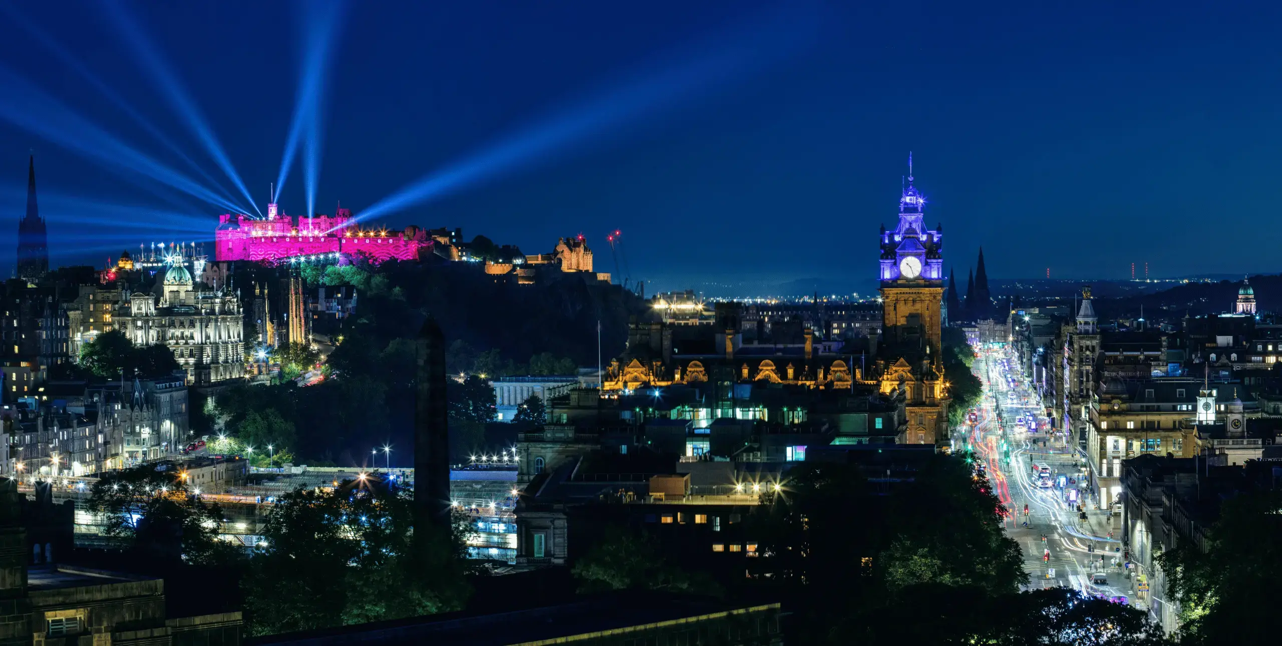 The Edinburgh skyline at night, with searchlights coming out of the castle on a hilltop, which is illuminated in pink light. A busy street is lit up to the right of the image.