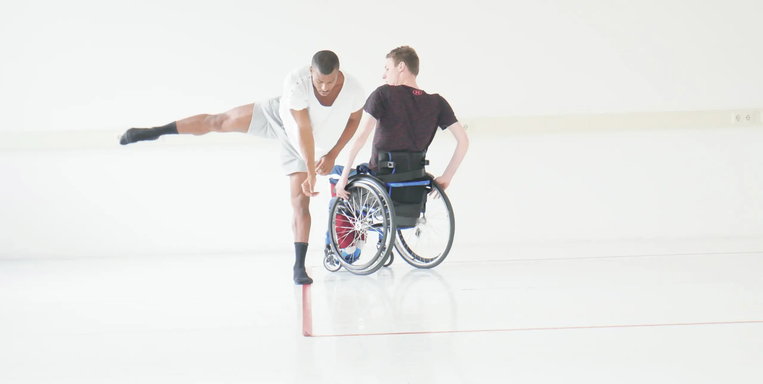 Two male performers stand next to each other in a white space. The man on the left stands in the foreground, bending forwards at the waist with his right leg outstretched. The performer behind and to his right uses a wheelchair and sits with his back to the camera.