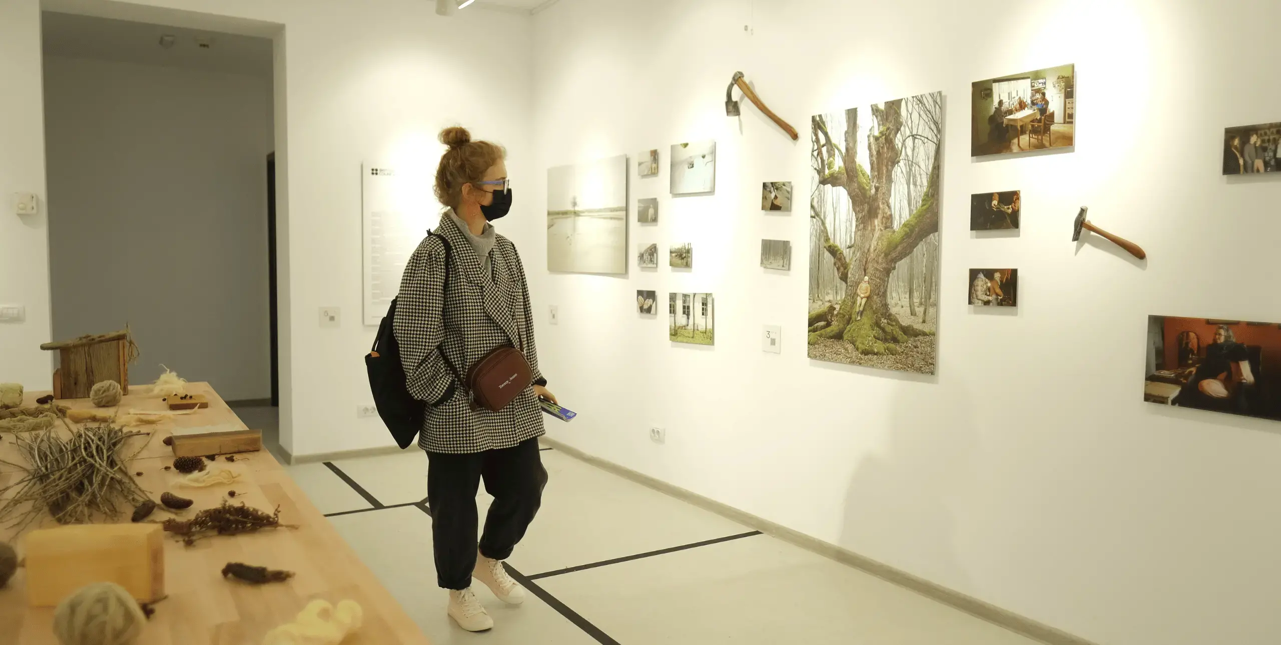 A woman in a face mask in a gallery space, looking at a wall-mounted display of tools and a painting of a tree.