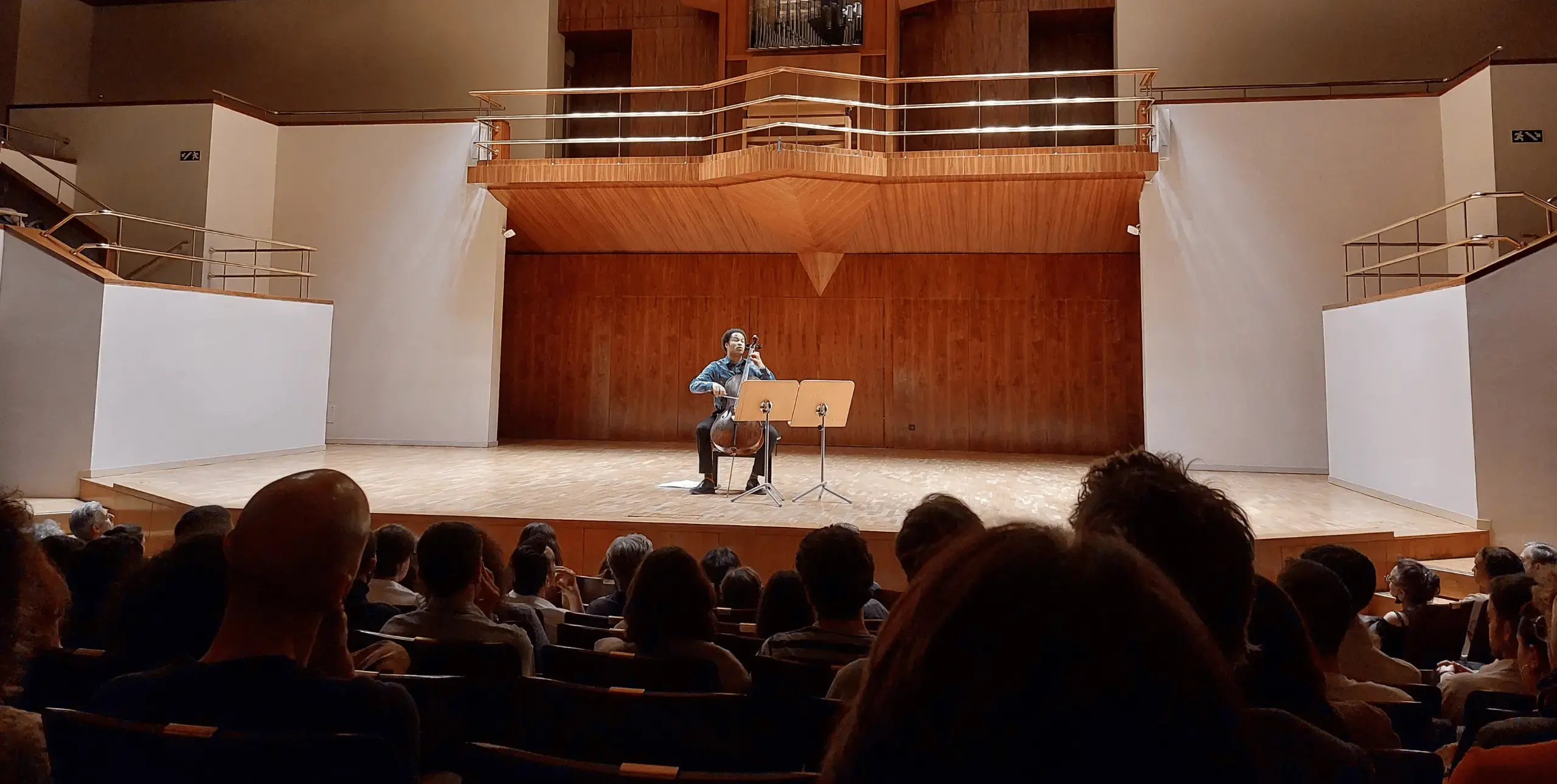 A large concert hall with modern chandeliers and a wooden stage. Sheku Kanneh-Mason is performing solo on the stage with a cello, while the audience sits attentively in the foreground, watching the performance.