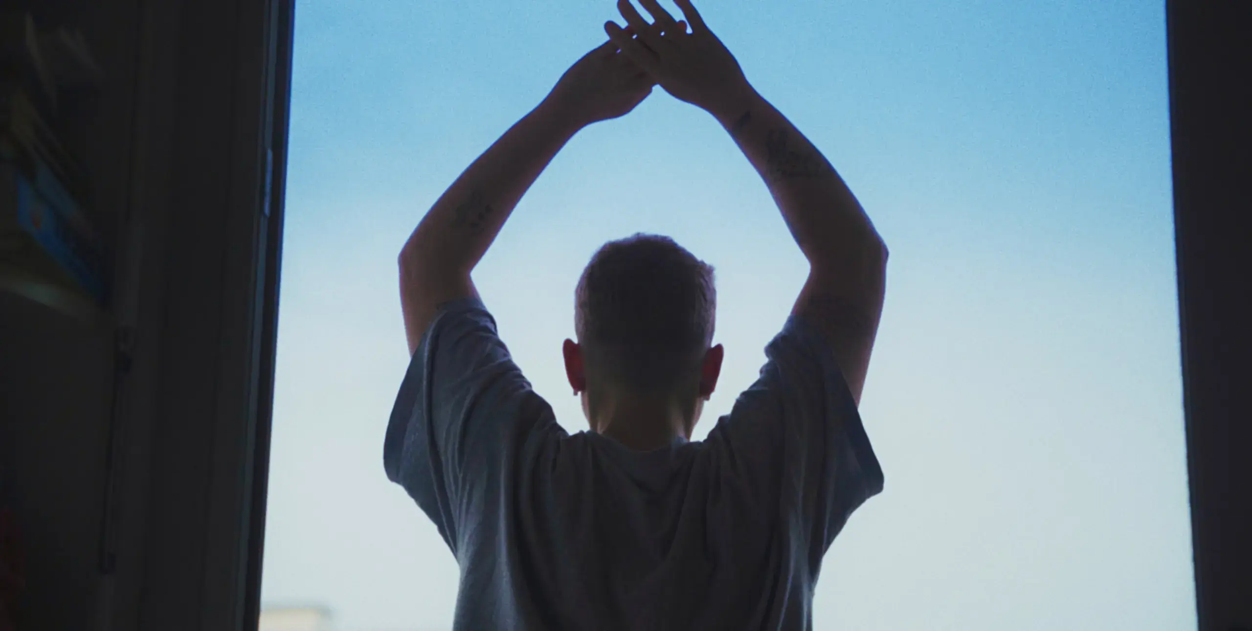 A young man stands in an open doorway, facing away from the viewer, silhouetted against a pale blue sky with his arms held up above his head, and his hands joined together.