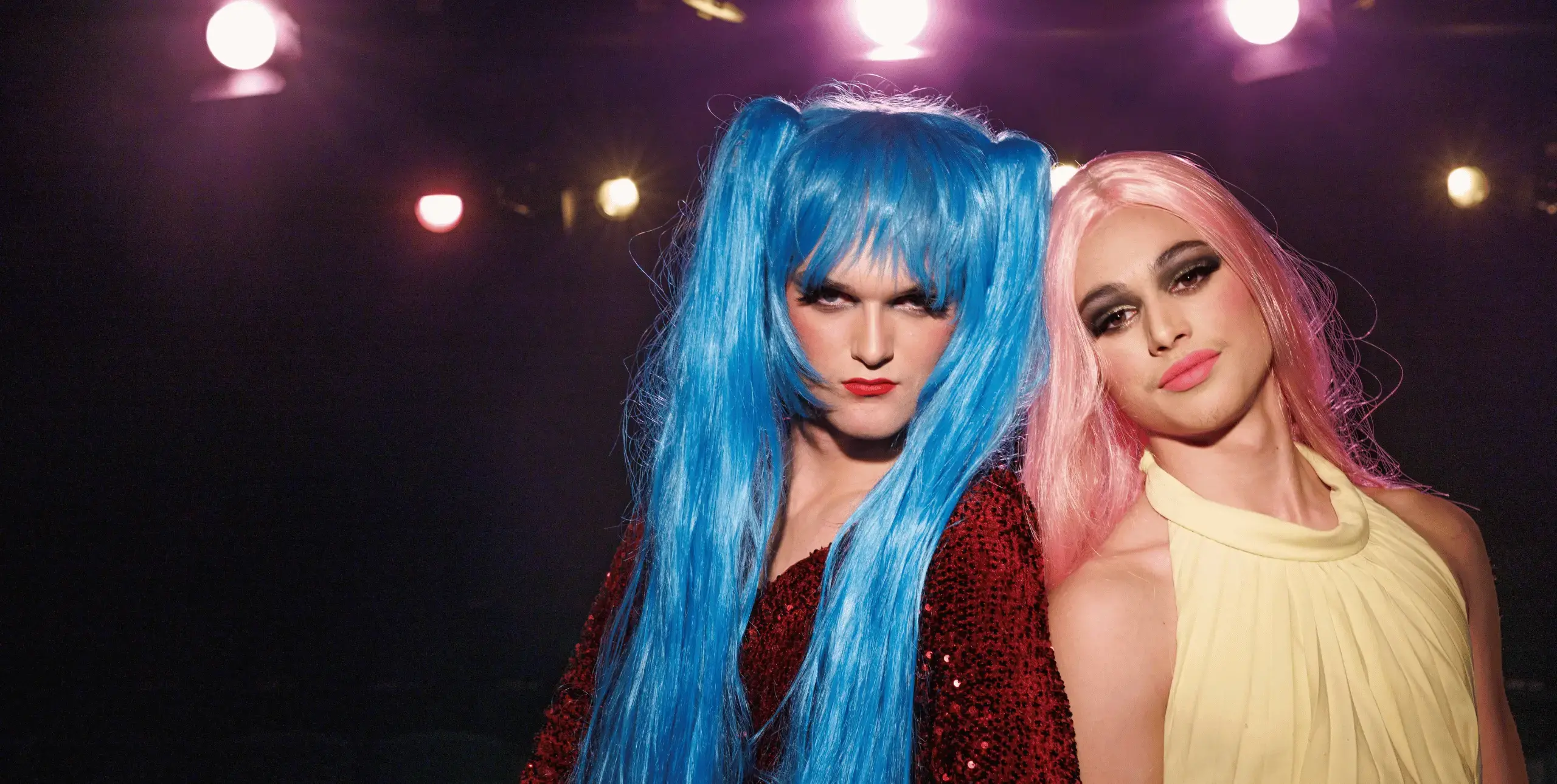 Two teenage boys dressed in drag stand side by side, shoulder to shoulder to the right of the frame. The boy on the left wears a bright blue wig in pigtails and the boy on the right wears a long pink wig. There are bright lights, resembling stage lighting, above their heads and they stand against a dark background.