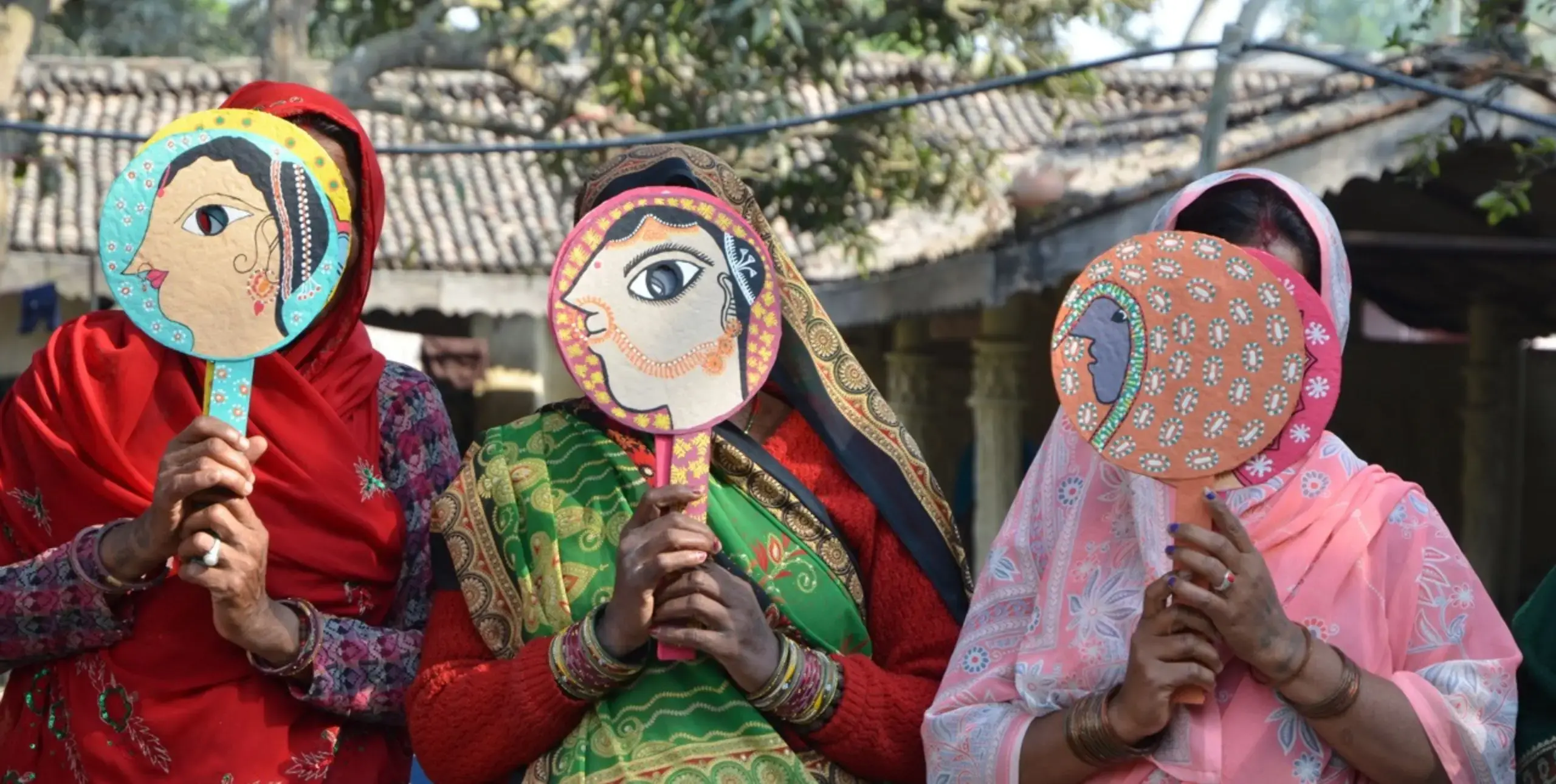 Three women in saris stand in a row with their faces obscured by hand-held masks on sticks with the faces of women painted on them.