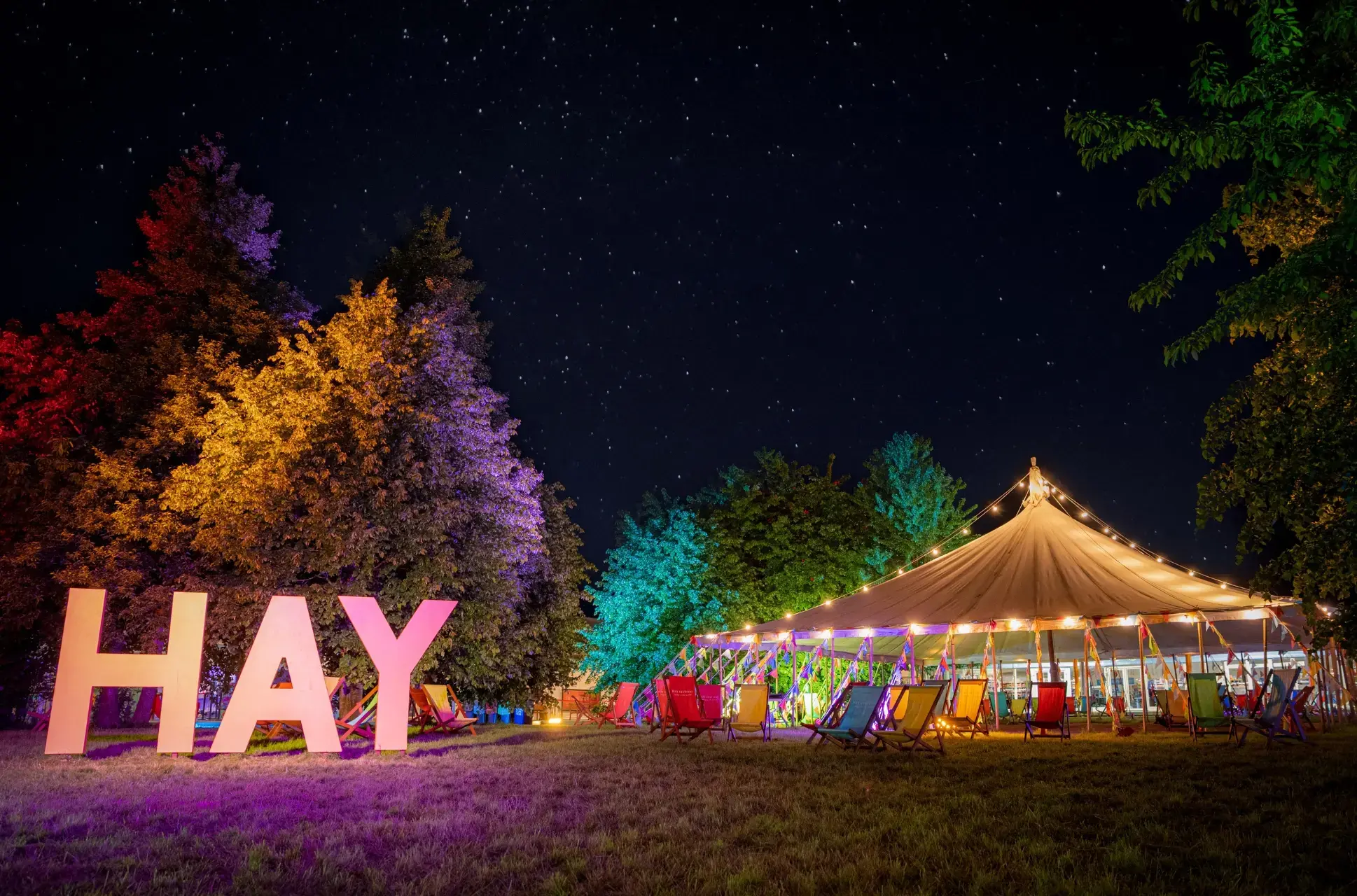 Hay festival scene at night an empty tent and colourful deckchairs next to big letters that say HAY