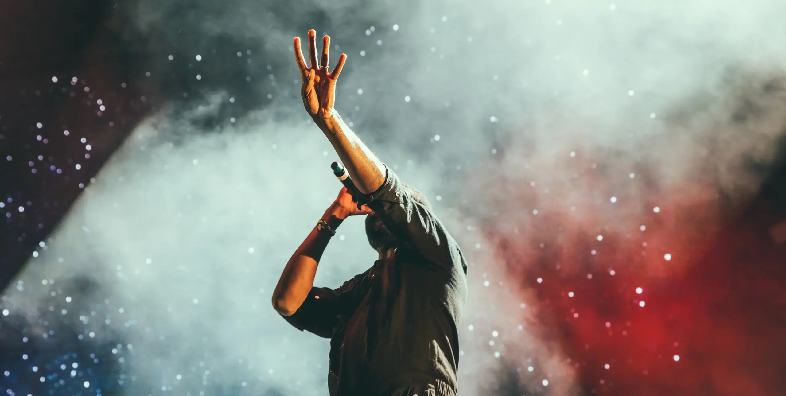 Man on stage with bright lights and white smoke, holding a microphone in one hand and raising four fingers into the air with the other.