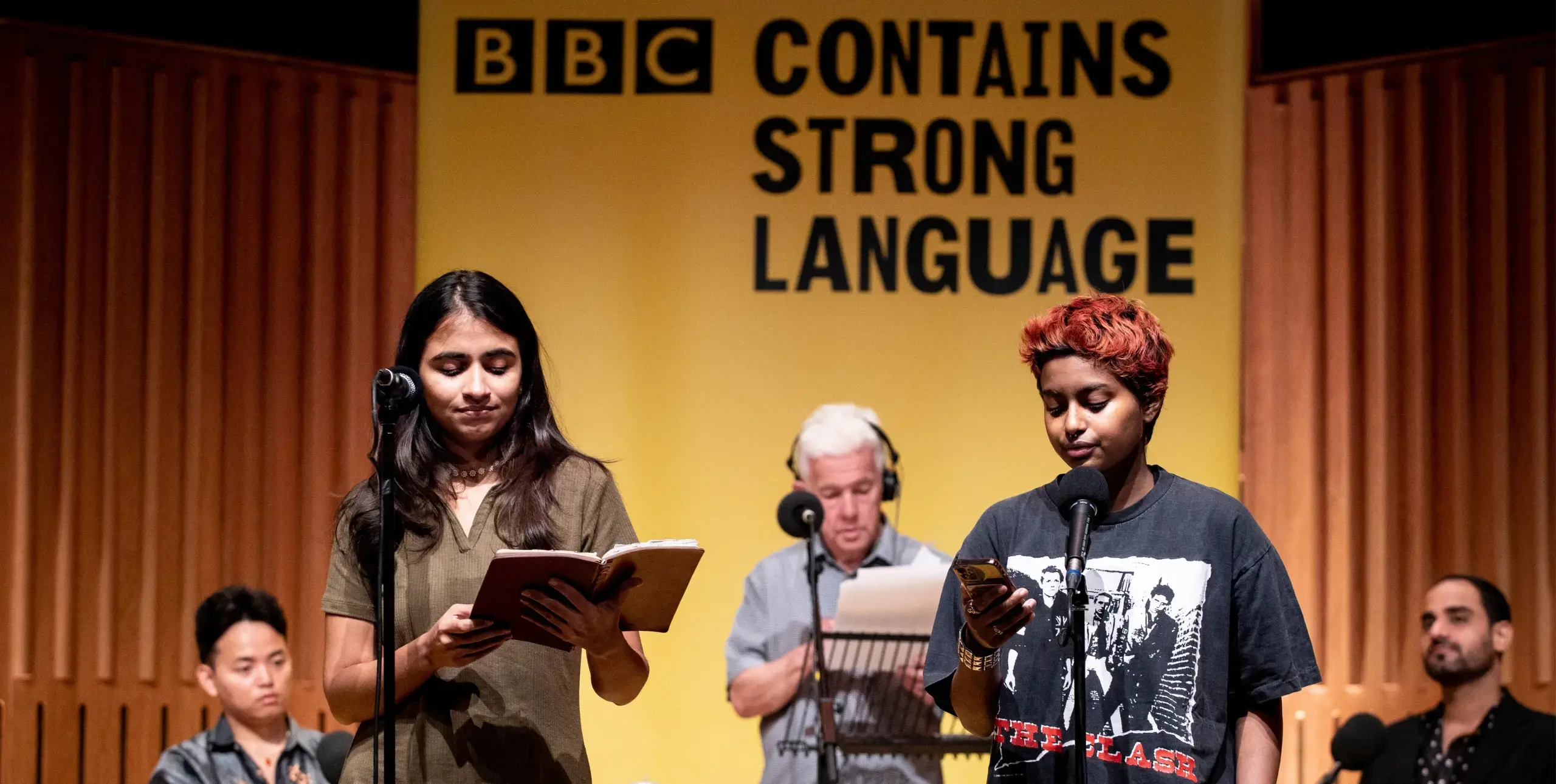 Two poets of Asian decent stand infront of a yellow banner at the BBC Contains Strong Language festival to perform their poems to a live audience.