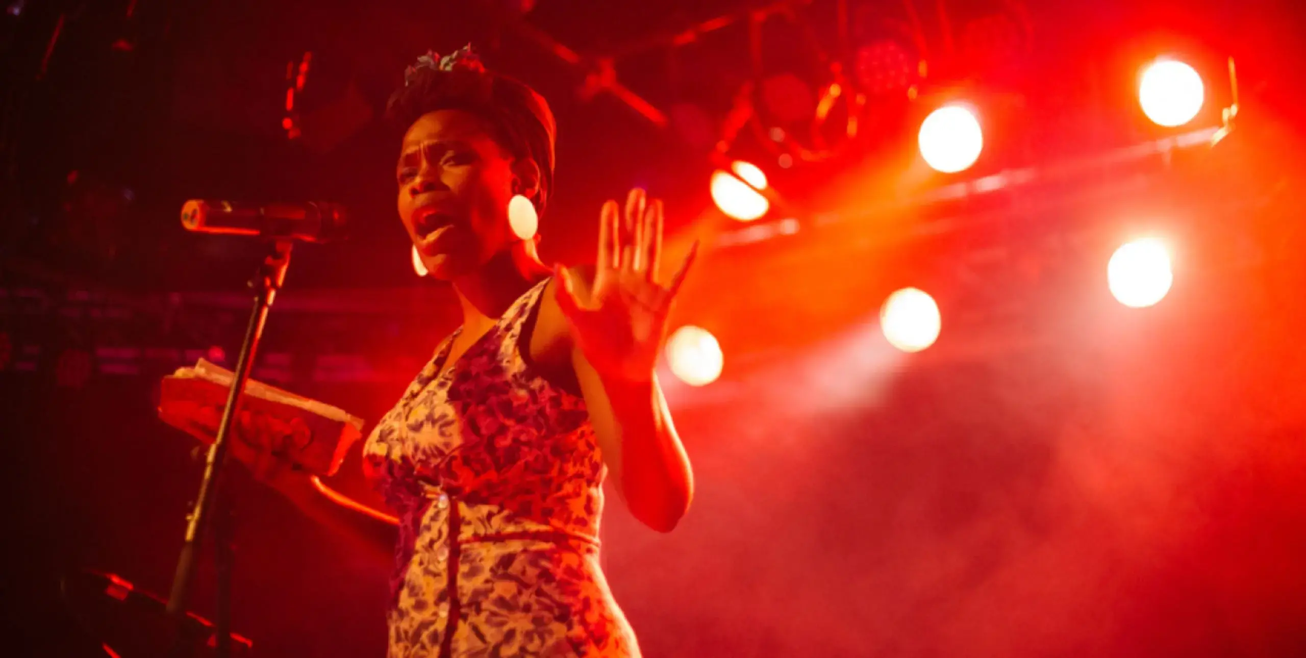 Vanessa Kisuule stands performing on stage infront of a microphone under red lights and smoke.