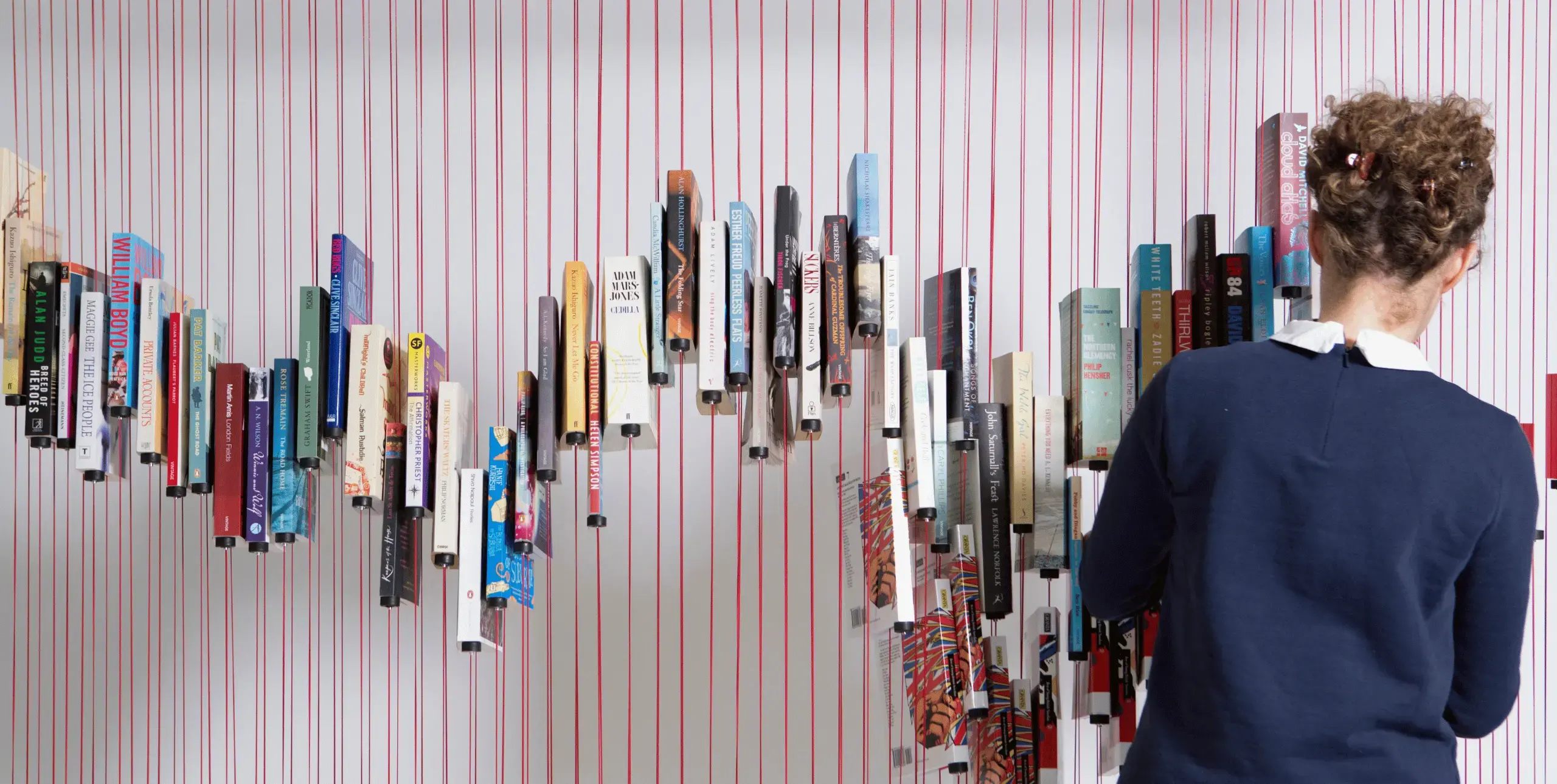 Paperback books suspended on vertical red strings at different heights, creating a zig-zag pattern. To the right of the image, a woman stands with her back to the camera, adding or removing books from the string ‘bookcase’