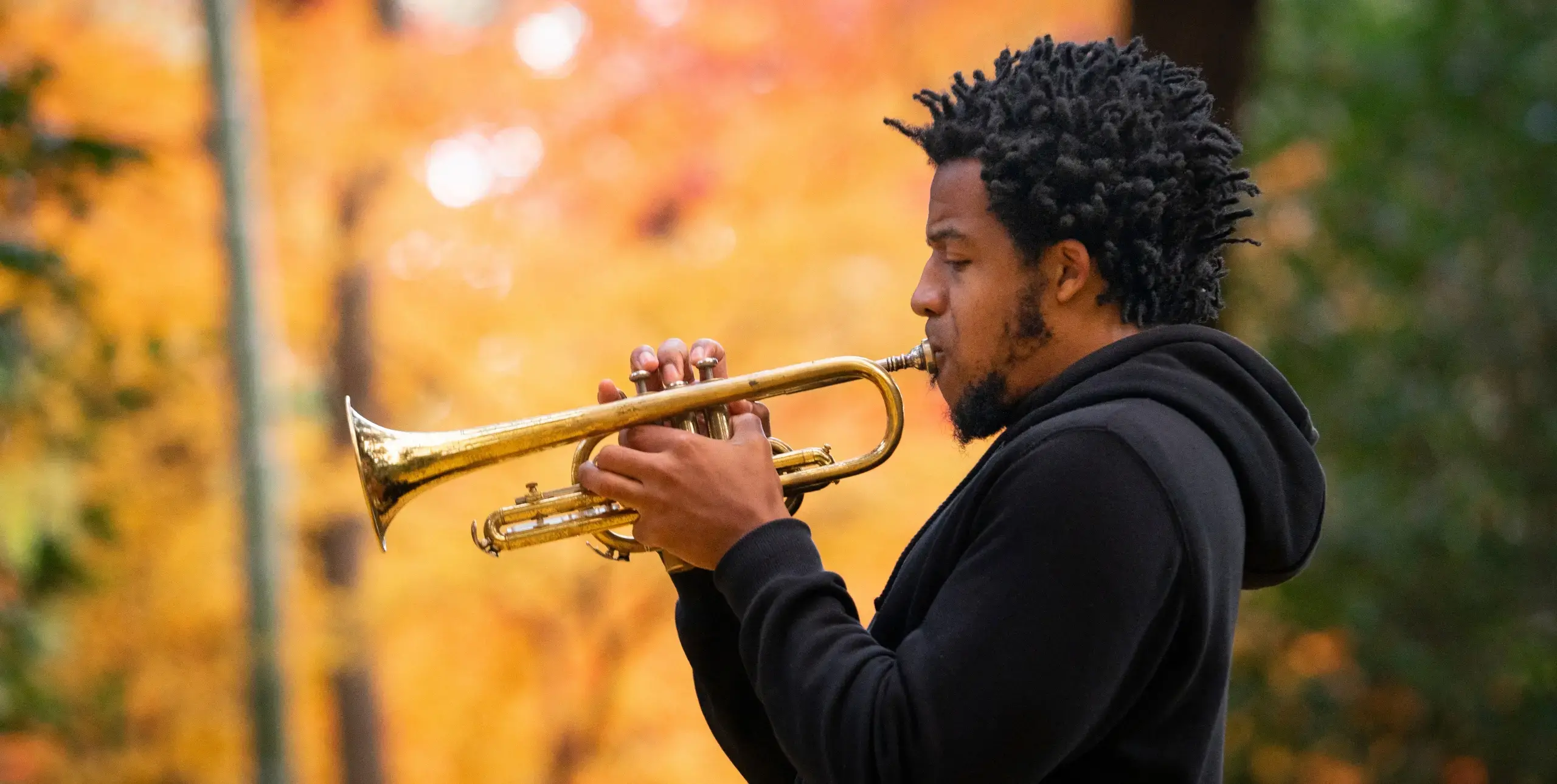 Man in black hoodie playing a trumpet in a forest.