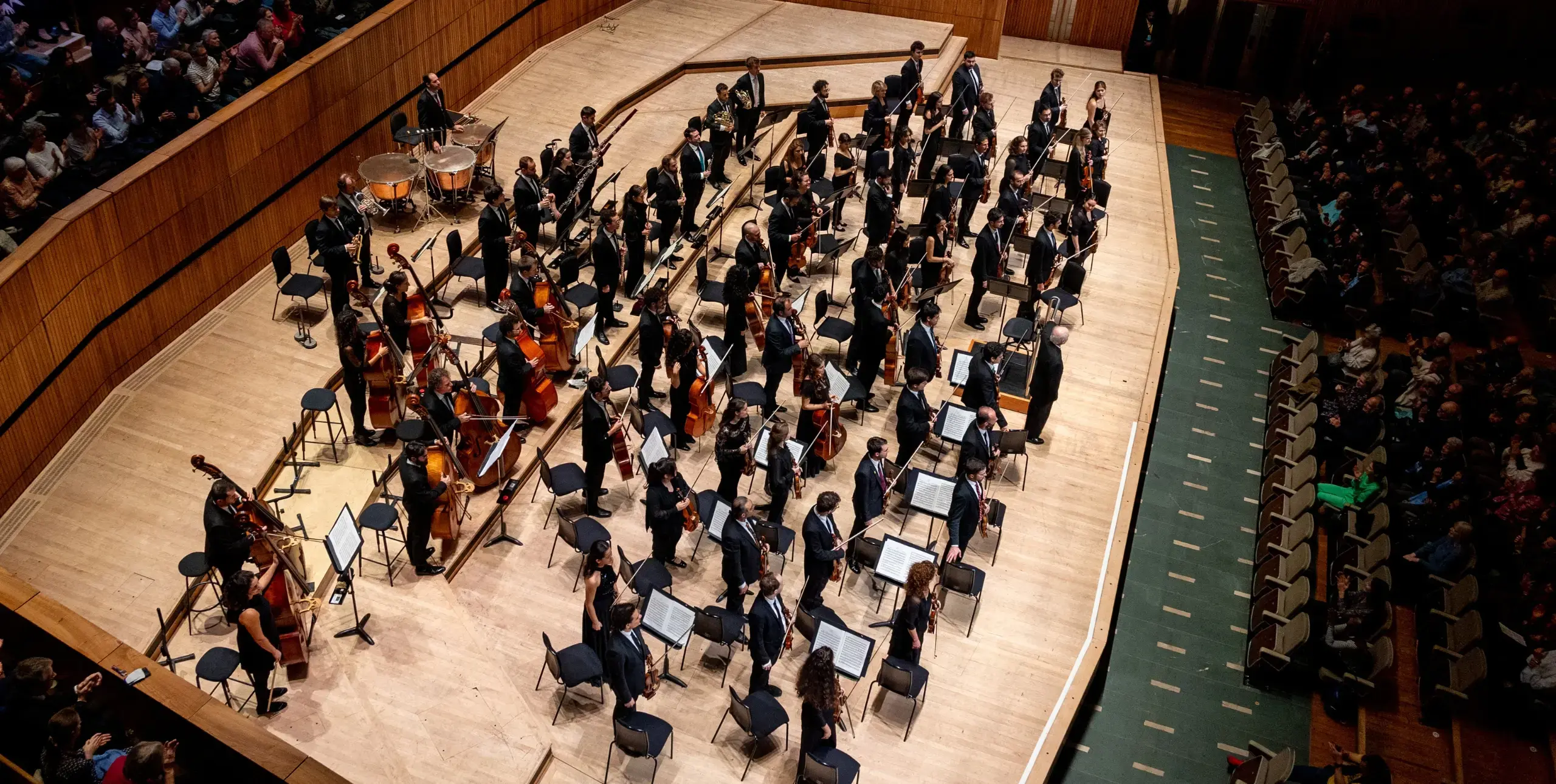 Classical orchestra ensemble, viewed from above, on stage with strings, woodwinds and percussion instruments.