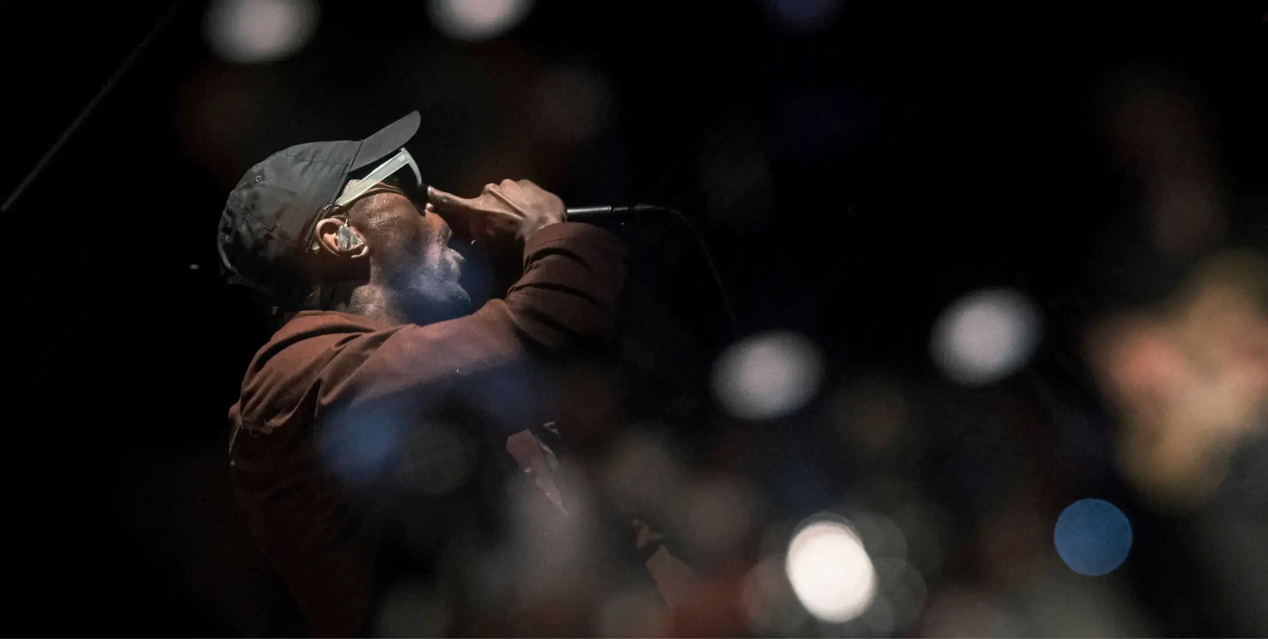 Man wearing cap and sunglasses, holding mic, with a dark background and blurred lights around him.