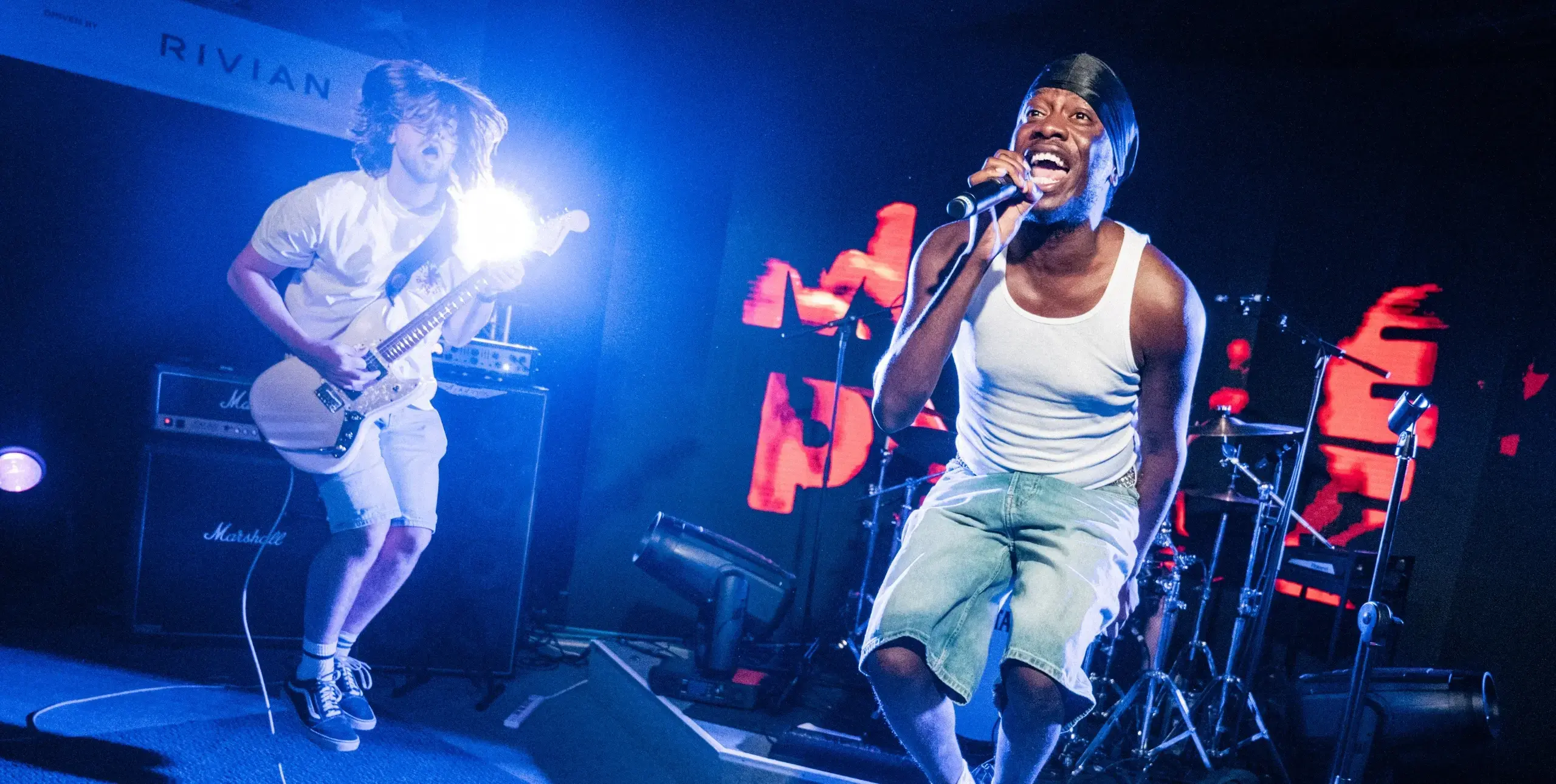 On stage, electric guitarist on left, jumping into the air; vocalist on right wearing white vest also jumping.