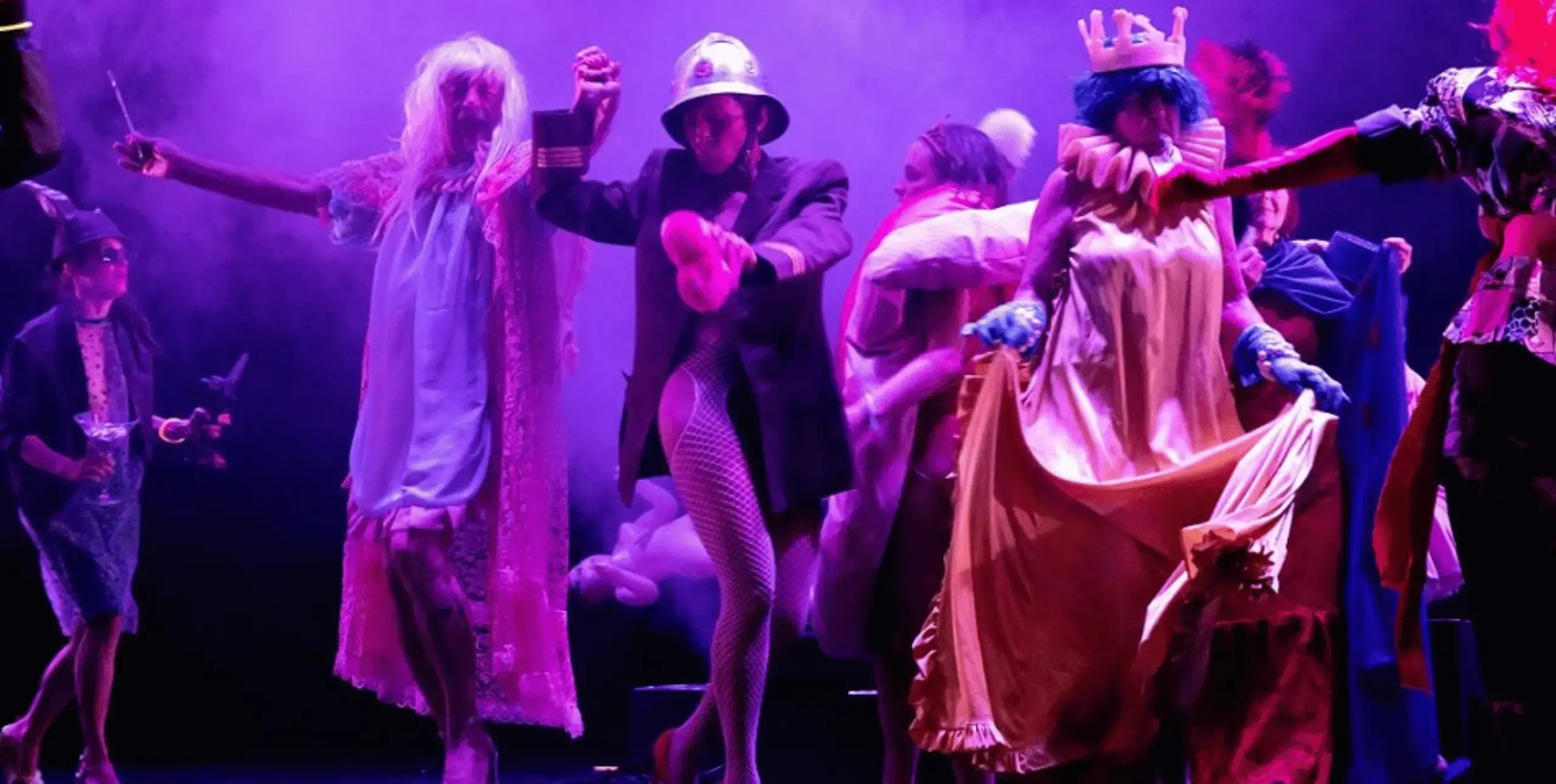 Premiere of ‘Nobody Whistles Anymore’ in Ljubljana, May 2023. Visual description: On the darkened stage a crowd of people in colourful costumes is dancing. At the front of the stage: a person in pink dress with a pink crown, a male in drag and a woman with metal helmet, military jacket and fishnet stockings. Flower petals on the floor and some smoke in the background.