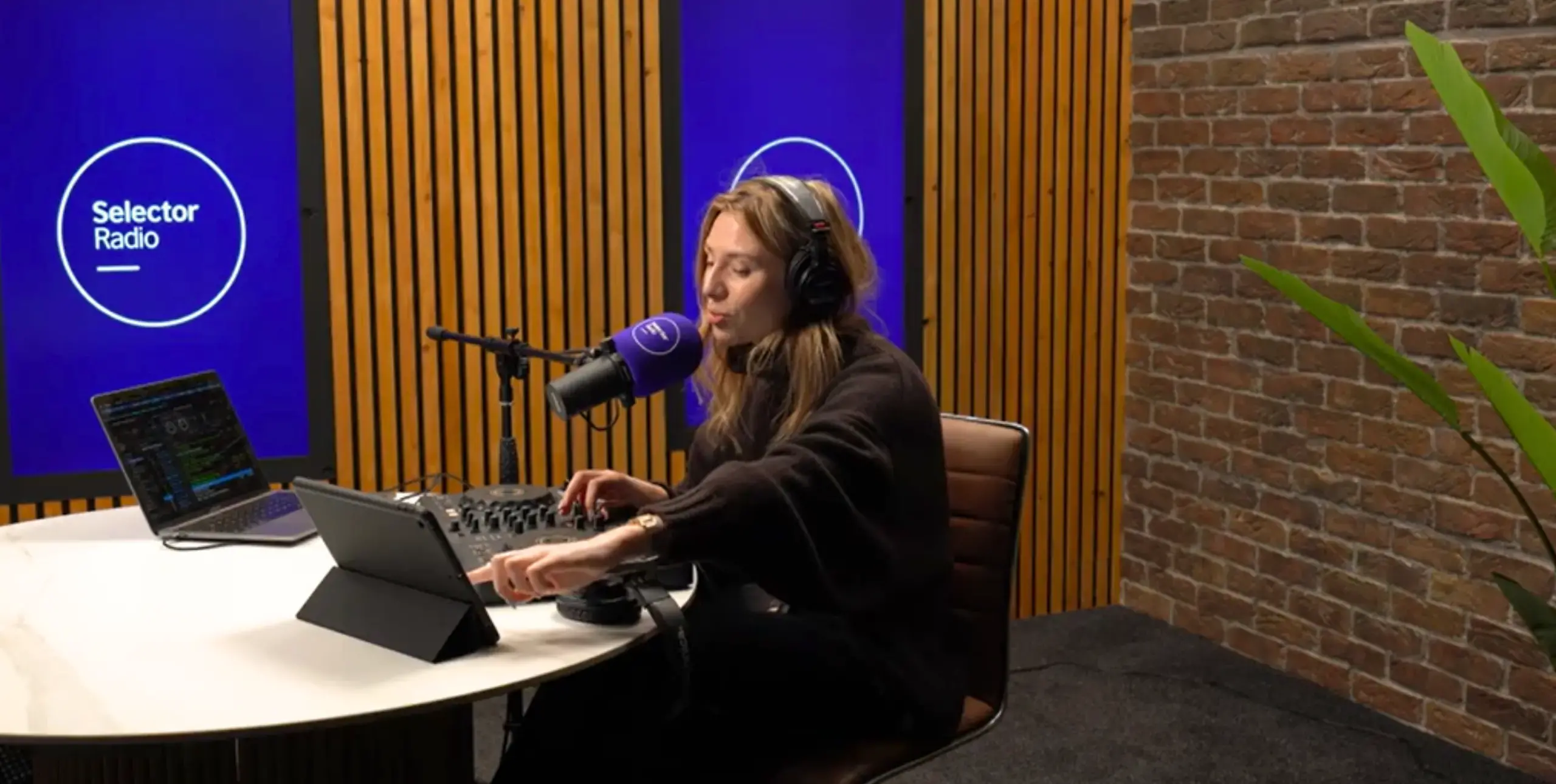 Lady in radio studio with headphones on, micrphone in front of her, at a desk, two laptops next to her, Selector Radio logo on a screen behind her.