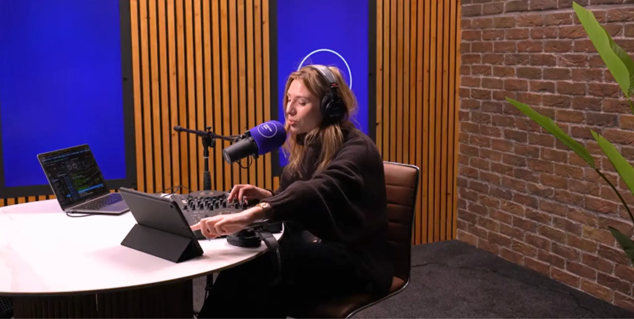 Lady in radio studio with headphones on, microphone in front of her, at a desk, two laptops next to her, blue screens behind her.