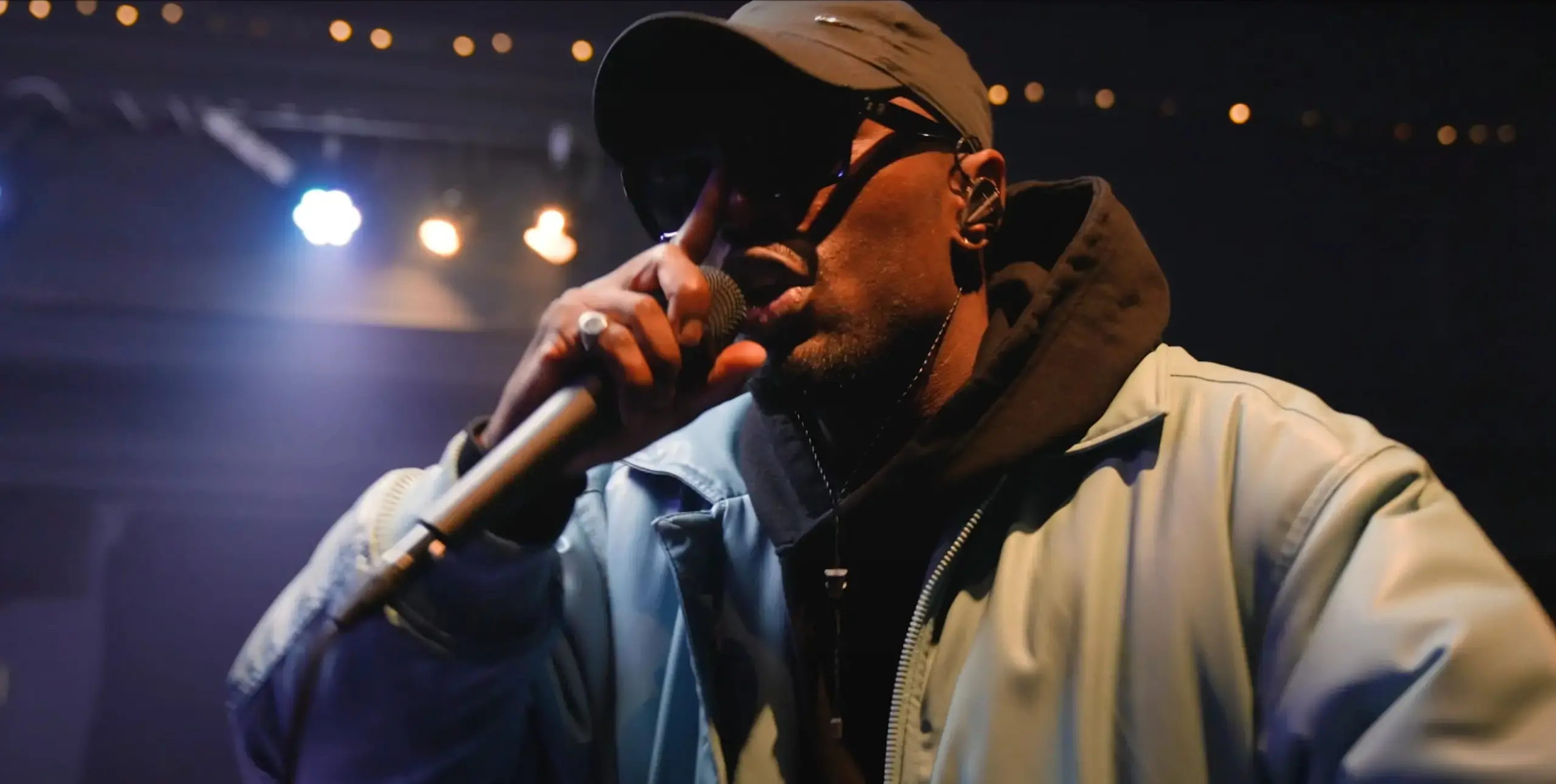 Close up of man, holding microphone to his mouth, wearing hooded top, cap and sunglasses. Bright indoor stage lights behind him.