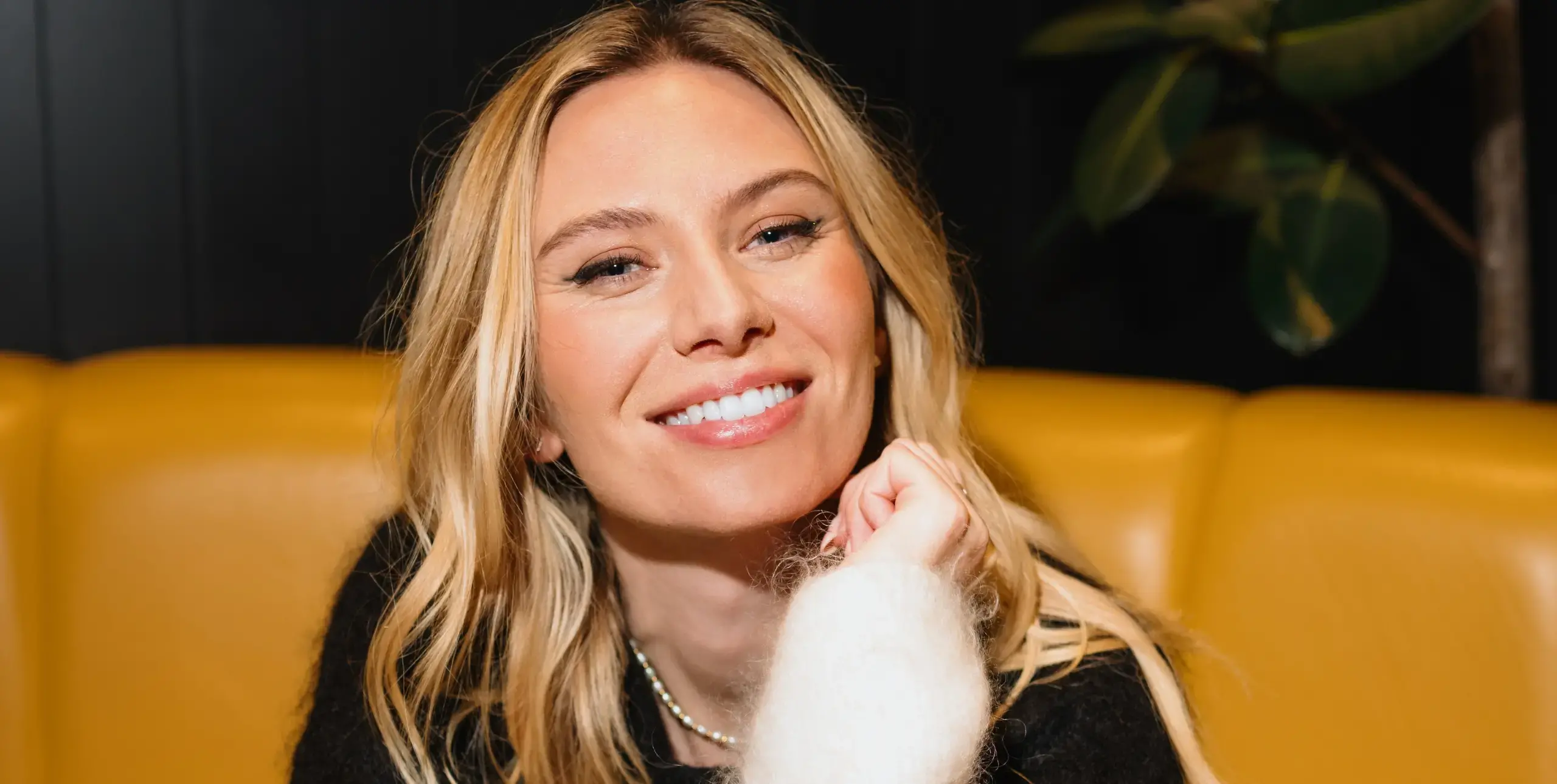 Close up of lady's face. She has blonde hair, sat on bright orange sofa, smiling.