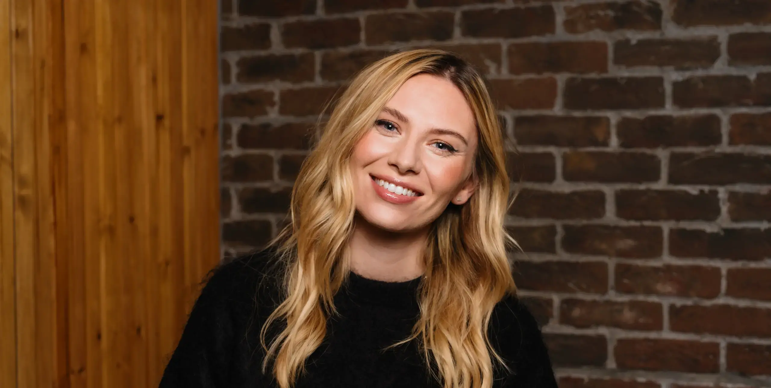 Lady with long blonde hair, smiling towards camera, in front of interior brick wall.