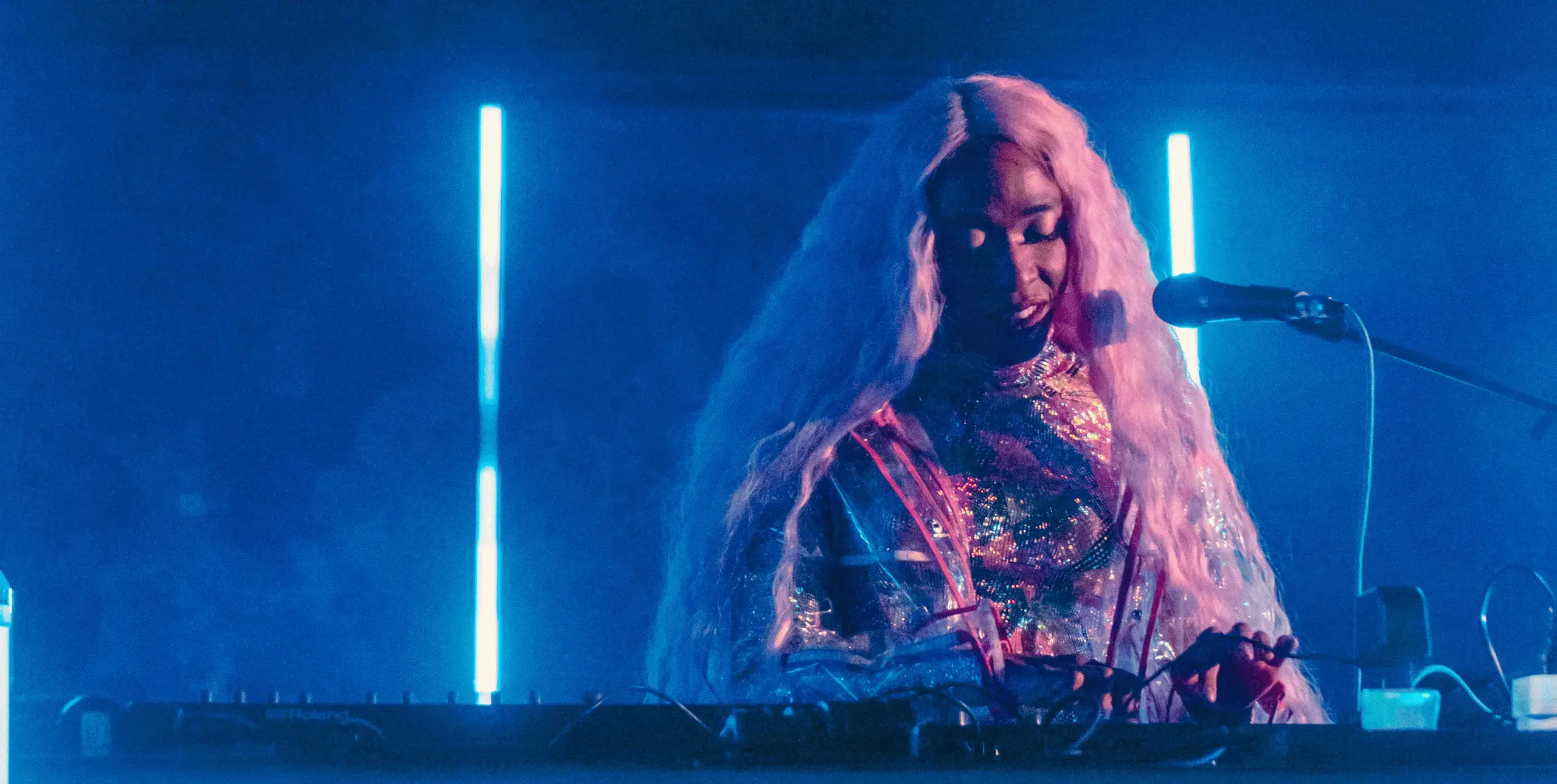 A Black woman, wearing a sequined outfit and with long pink hair, stands in front of a microphone and operates a sound desk. Behind her is a haze of dry ice and bright blue neon lights.