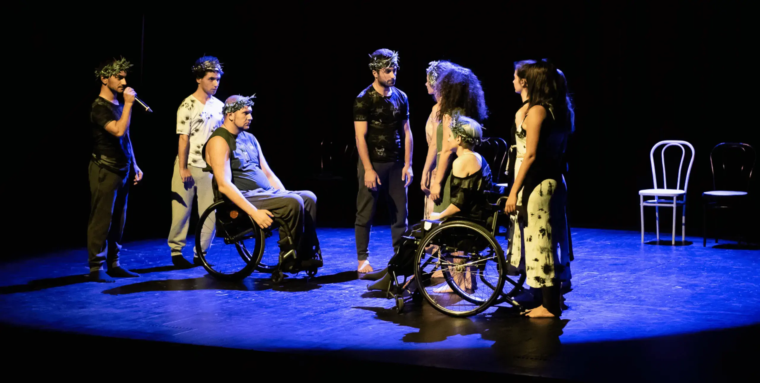 A group of performers, some able-bodied and some using wheelchairs, stand on a dark stage bathed in blue light. A white table and chair stand in the background-right of the image.