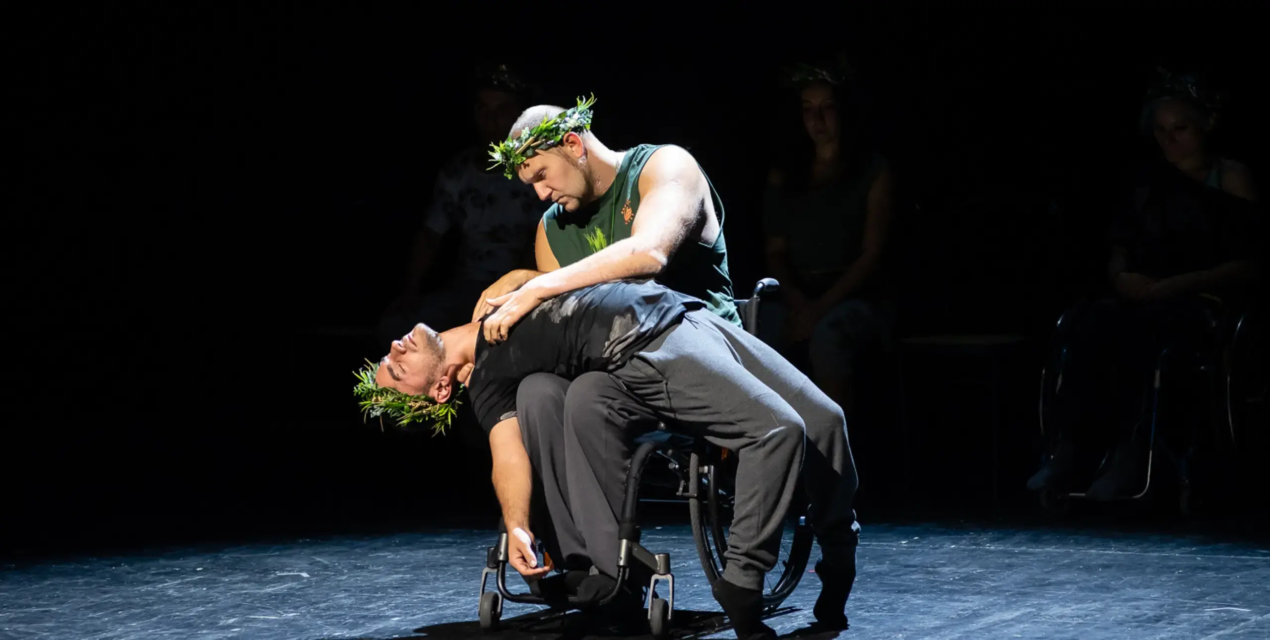 Two men on stage wear grass crowns and black costumes  in a dress rehearsal of Unlimited: Making the Right Moves.. One performer is in a wheelchair and the second performer bends his back over his knees in a dynamic back bend.