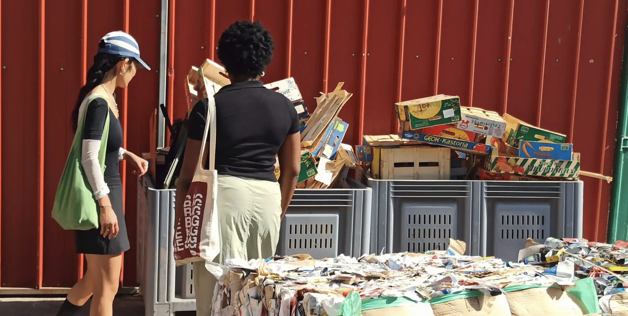 Two women stand with their backs to the camera, looking at a recycling unit which is overflowing with wooden crates and cardboard boxes. Cubes of compacted materials for recycling stand in the foreground, slightly out of focus