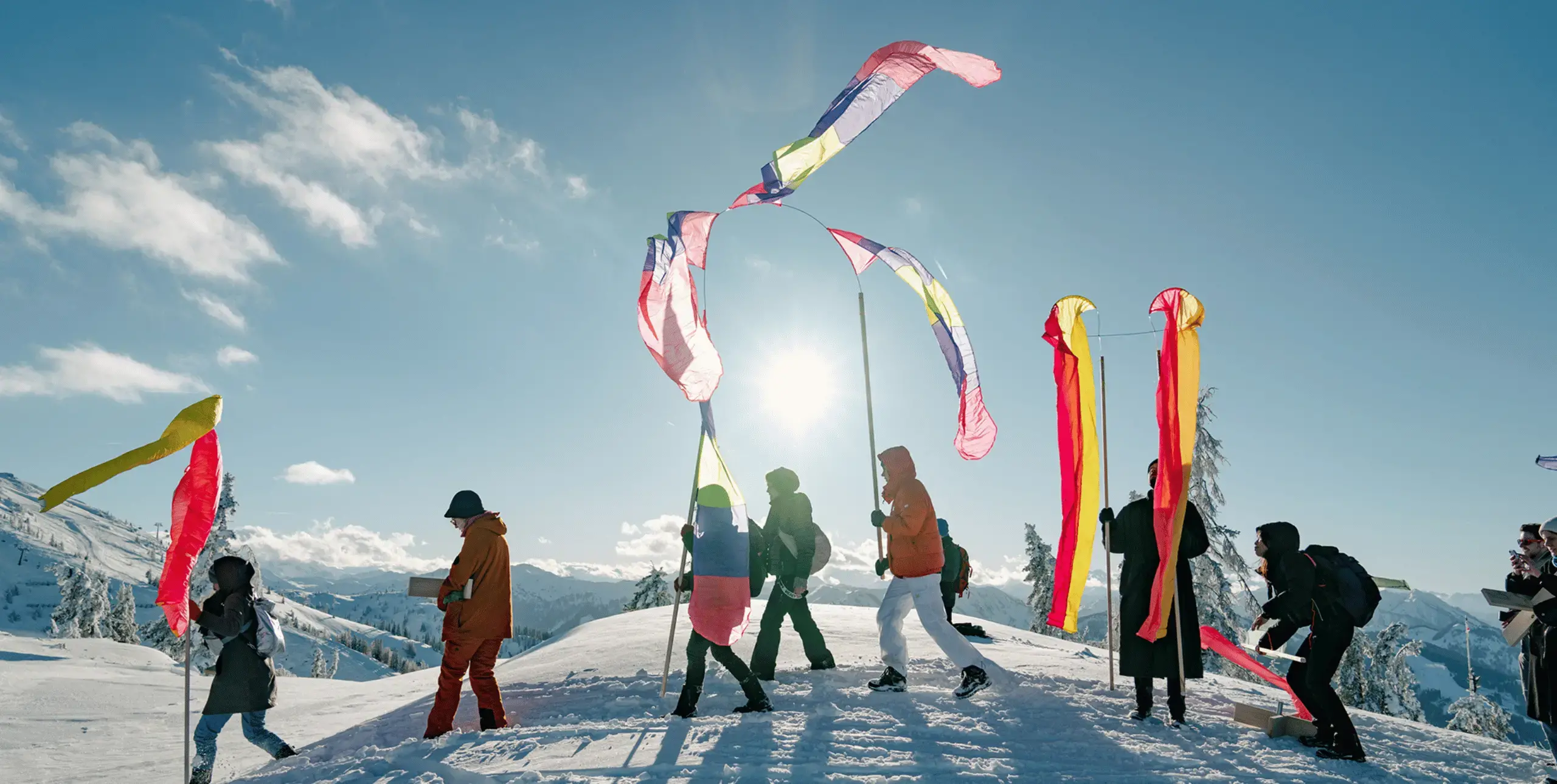 Eight figures walking across over snowy ground from right to left of the frame. They are wearing clothing for cold weather and are carrying colourful banners or flags on long poles.The sky is blue with a few small clouds and the sun is in the centre-background above the figures' heads.