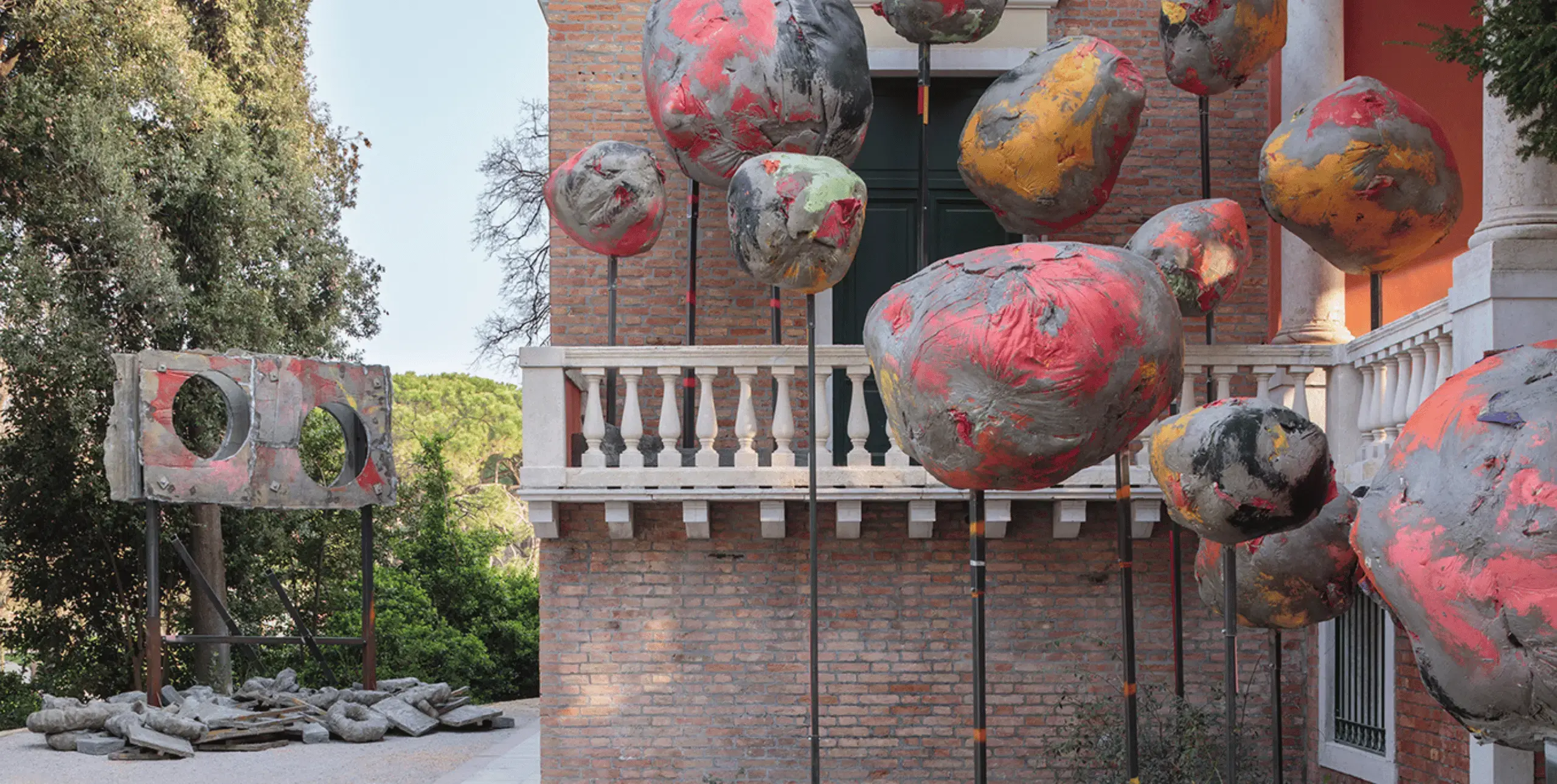 Large spherical papier mache sculptures of various sizes painted red, yellow and grey, mounted on sticks on the right/foreground of the image. Behind the sculptures the side of a redbrick building with a white balustrade can be seen. In the background on the left of the image stands another grey, red and yellow sculpture, which is shaped like a large placard mounted on two sticks, with two large circular holes cut into it. 