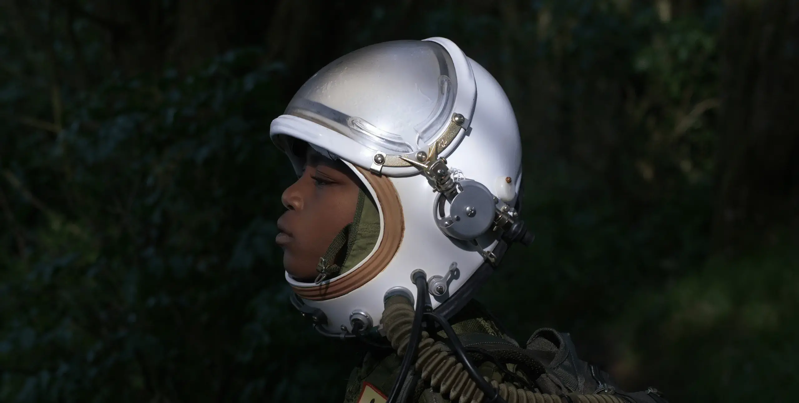 Still image from 16:9 video. Head and shoulders profile of a young Black child wearing a white space helmet against a dark green forest background.