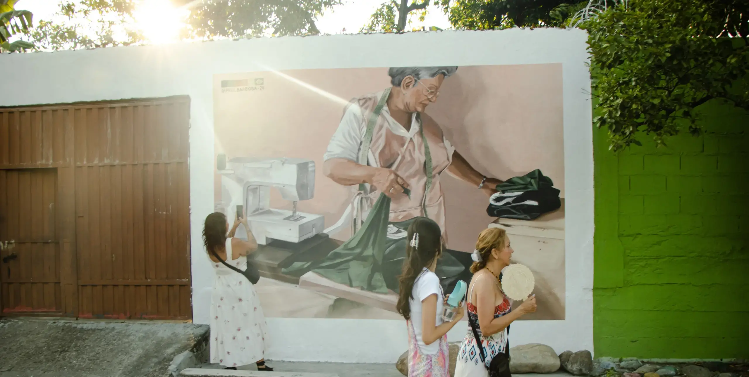 A painted mural shows a woman doing laundry on an outdoor wall. The sunsets behind the wall and passerbys look and take pictures.