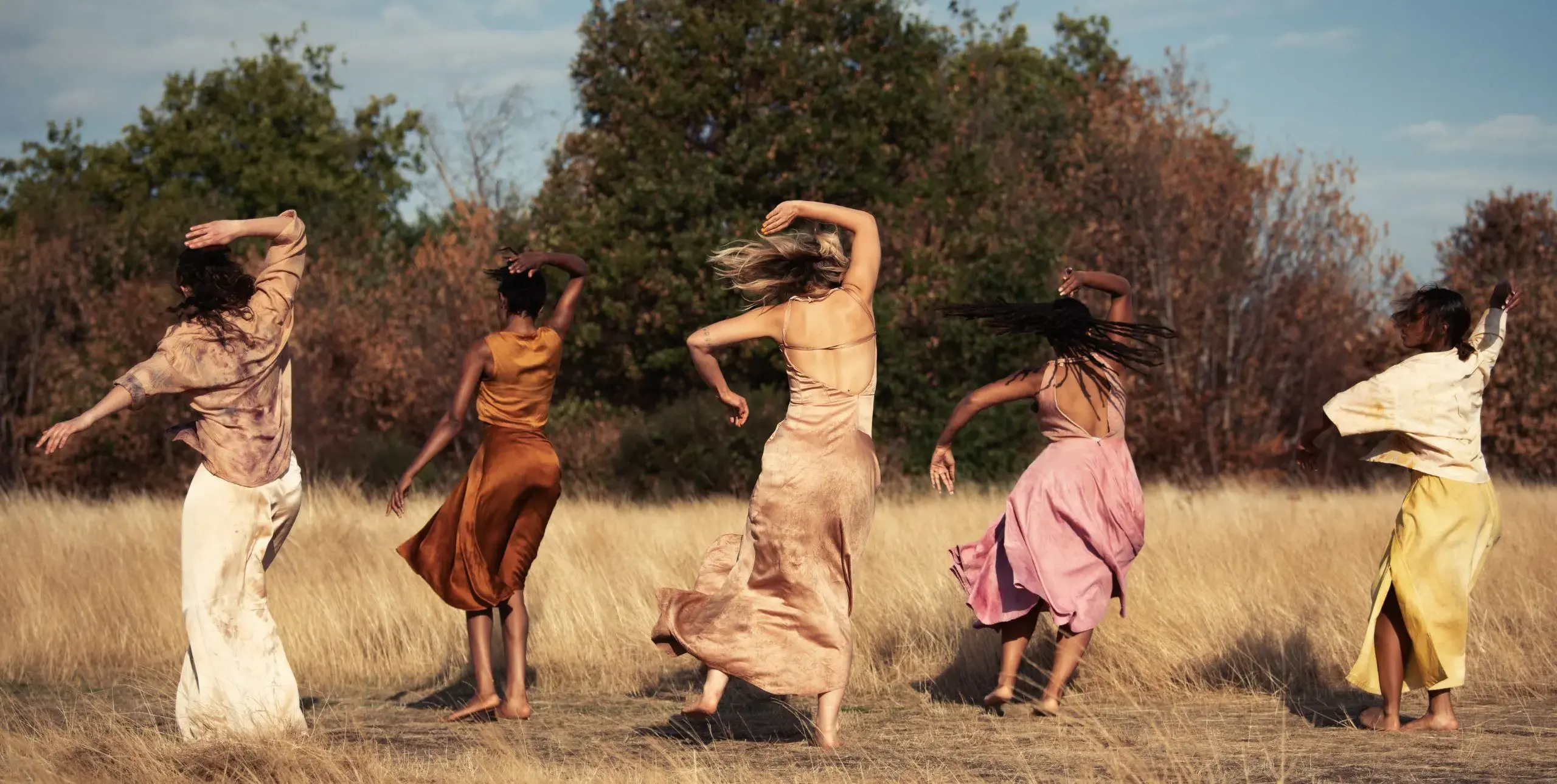 Five women in flowing dresses dance outside in nature