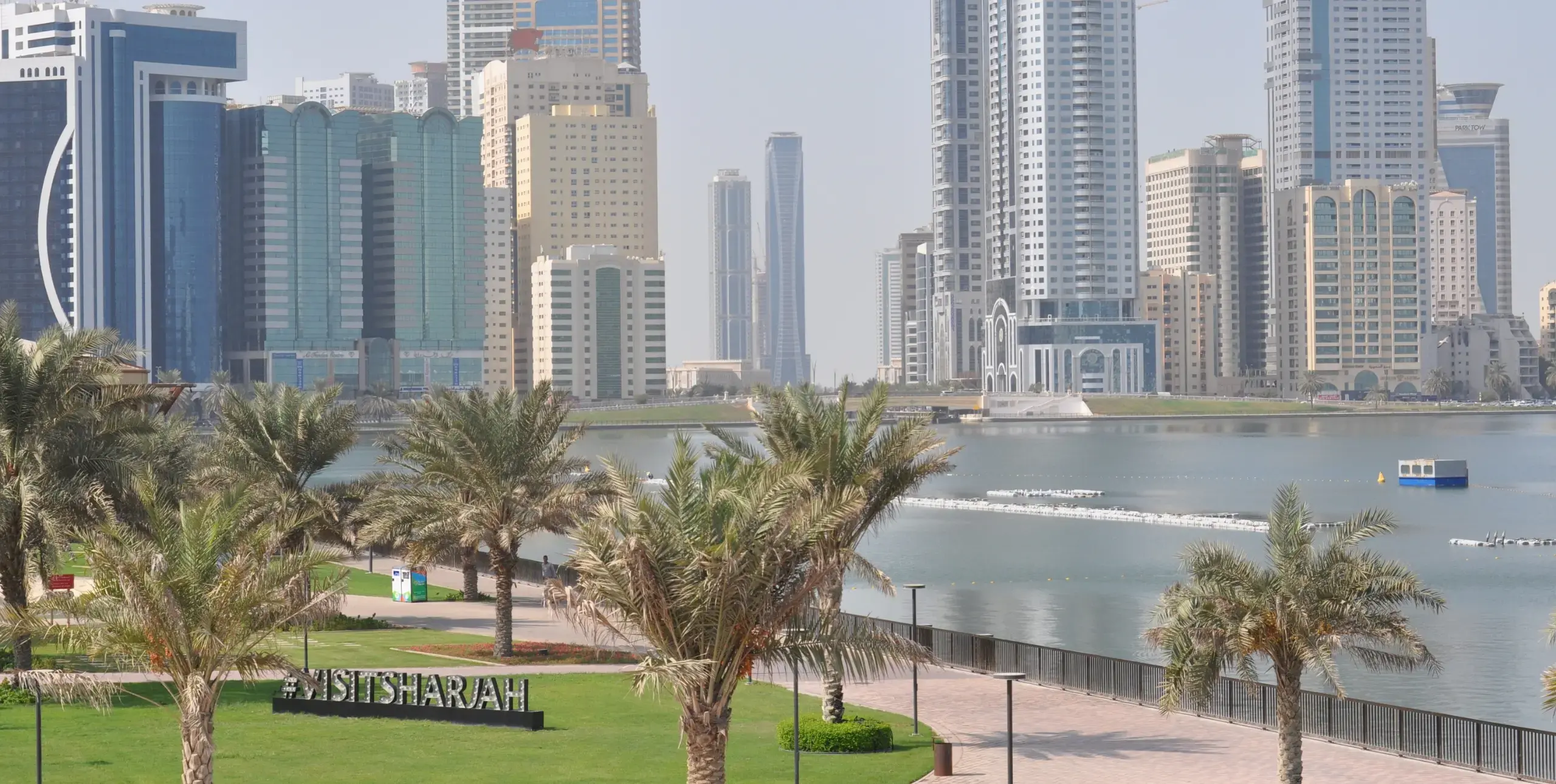 A waterfront view of Sharjah’s modern skyline, with palm-lined promenades in the foreground, sparkling calm water and high-rise buildings behind, and a “#VISITSHARJAH” sign nestled among the palms.