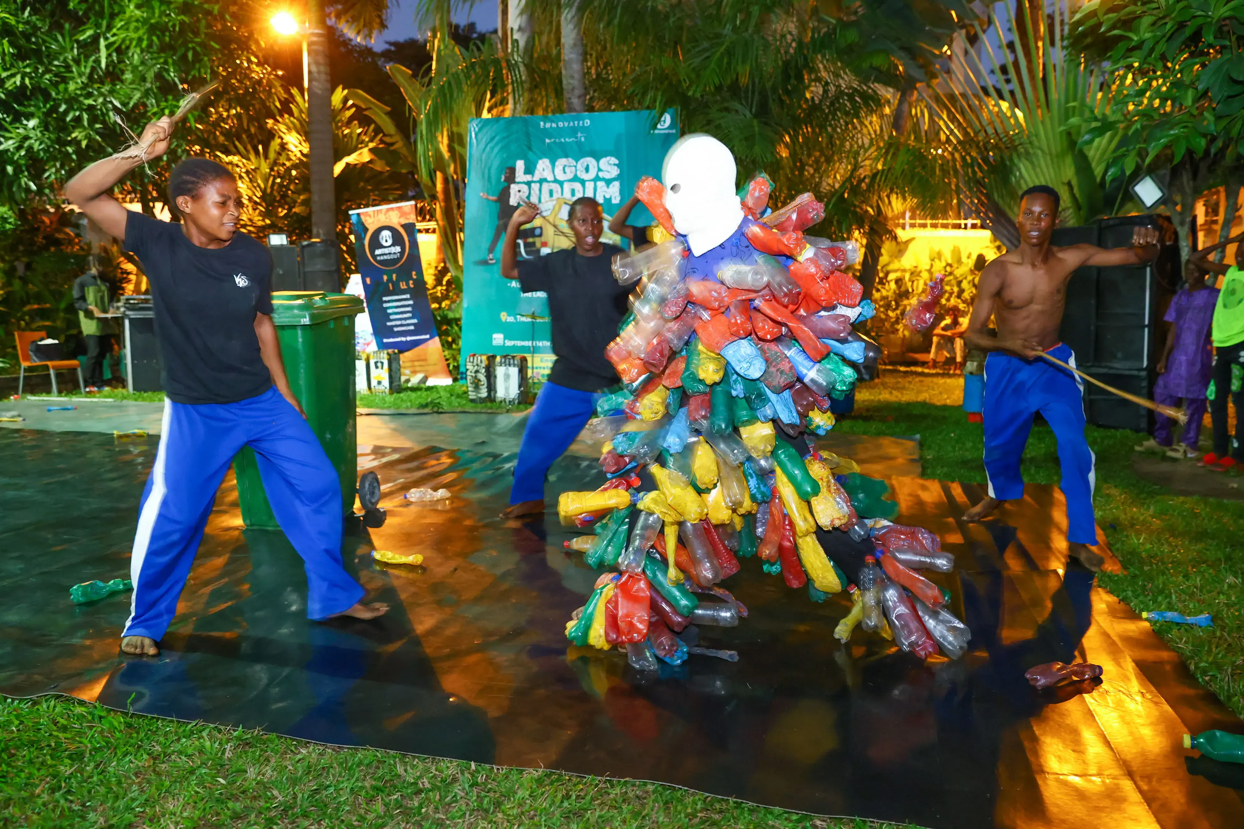 Outdoor performance featuring three performers surrounding a figure in a costume made from colourful recycled plastic bottles, with a wheelie bin and scattered plastic bottles on a stage beneath tropical trees.
