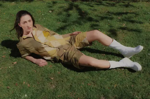 Fashion Revolutionaries - A female model lies on green grass, wearing an outfit of patchworked shorts and shirt in greens, browns and yellows. The model also wears white calf-length boots. 