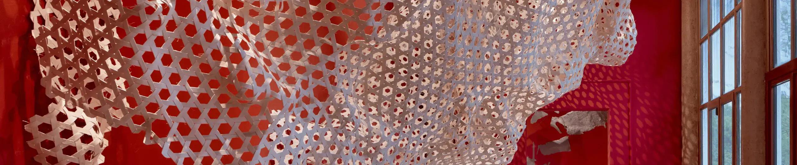 A large rattan weave sculpture hangs from the ceiling of the British Pavilion at the Venice Biennale. The windows of the Pavilion are visible to the right of the image, and the walls are painted red..