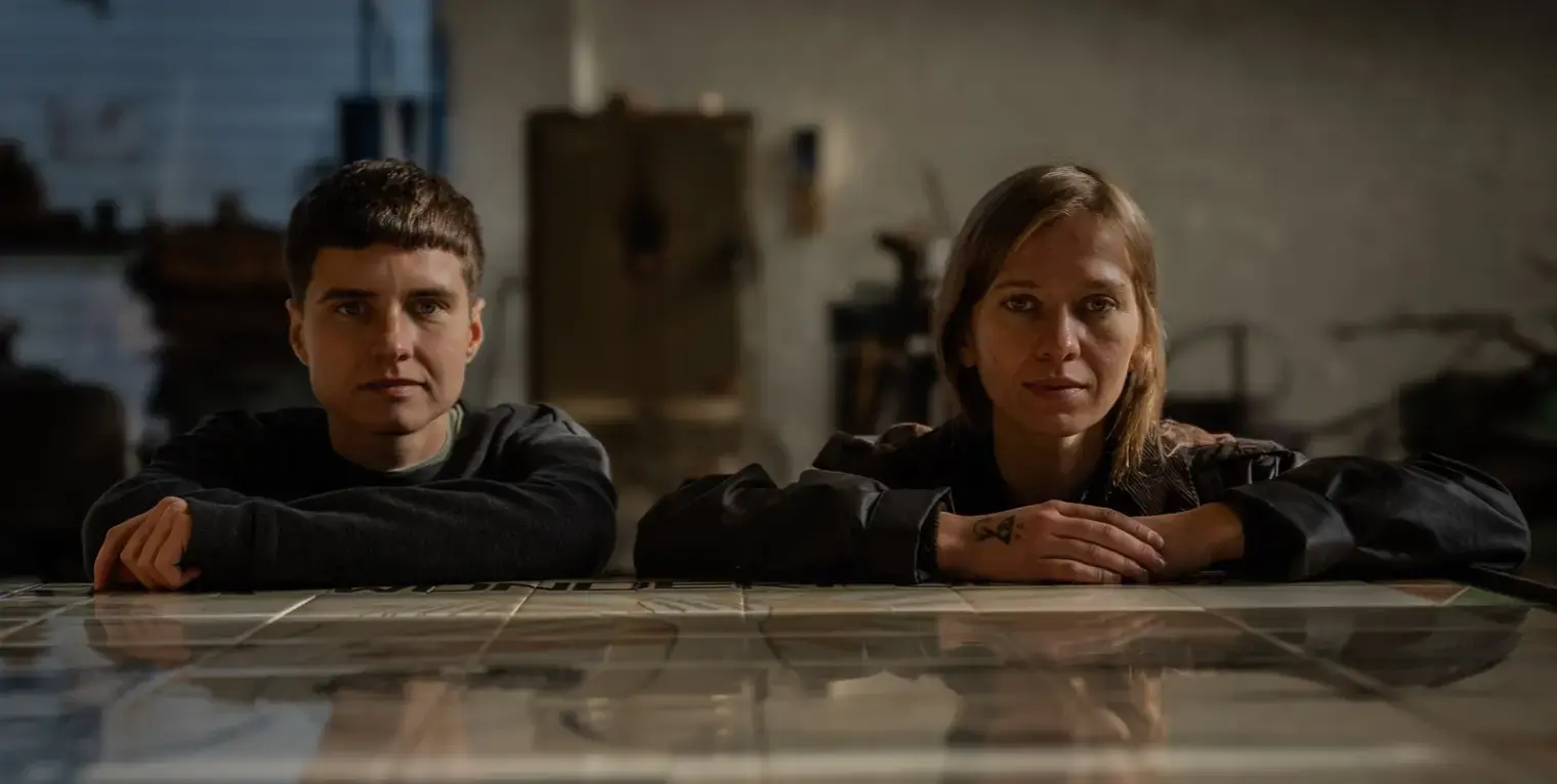 Two people sit alongside each other, staring straight at the camera, with their arms resting on a shiny tabletop.